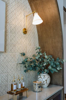 A brass sconce illuminates patterned tiles, foliage, and decorative bottles.