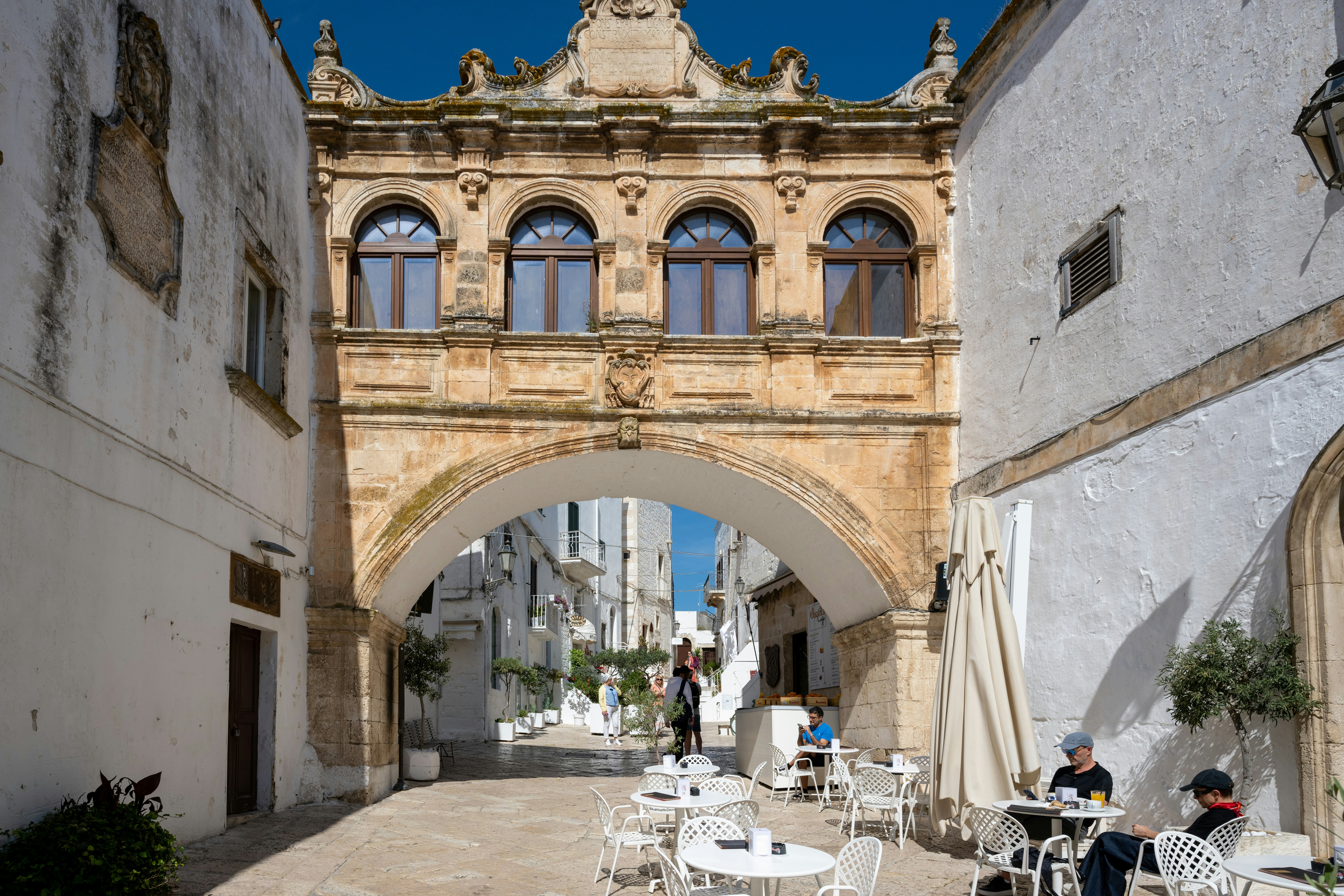 Un arco storico collega edifici bianchi sopra un caffè all'aperto.