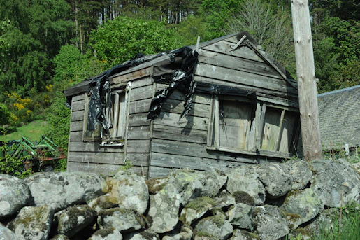 Weathered wooden shed behind a stone wall near a forest.