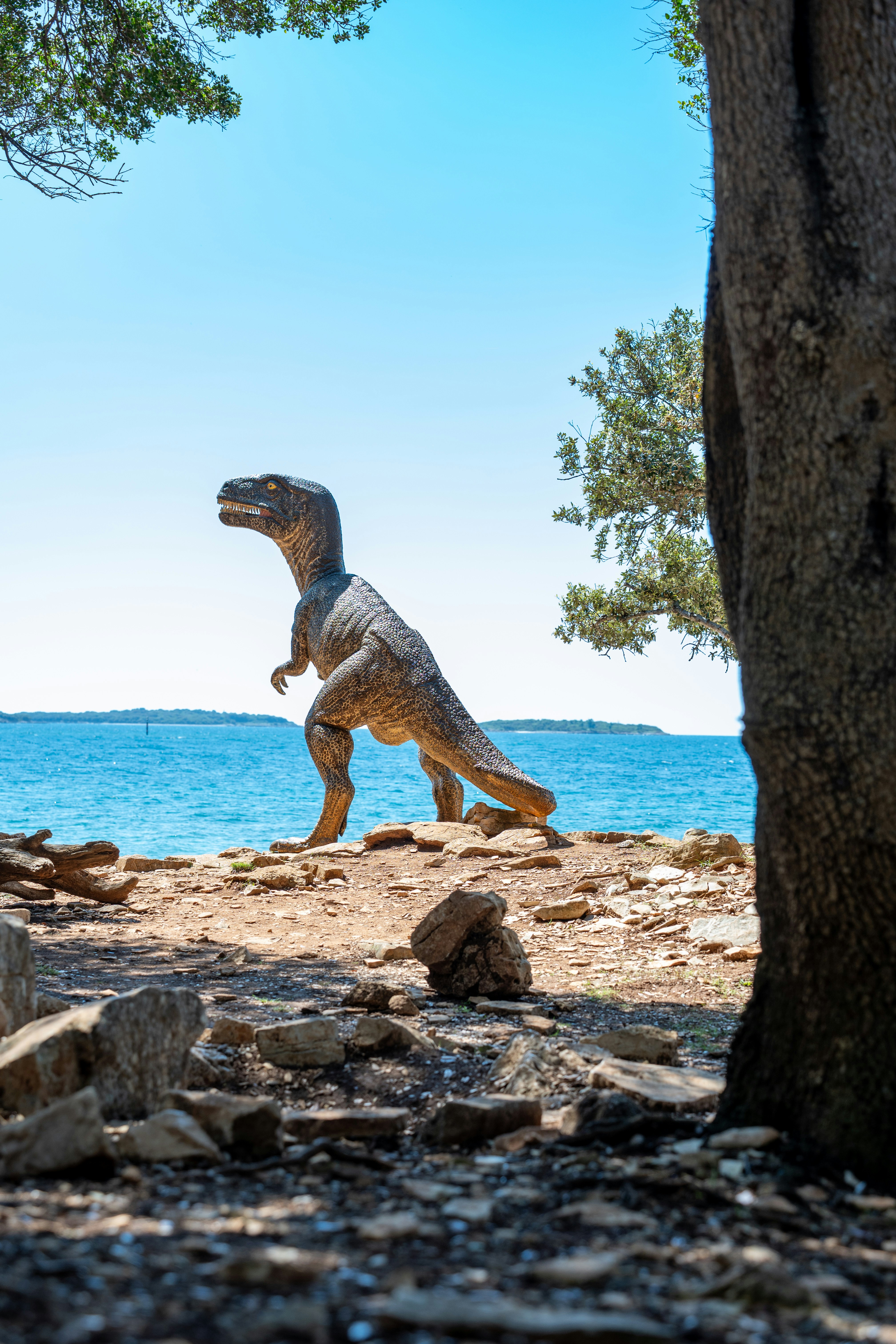 Patung dinosaurus di pantai berbatu di bawah langit biru jernih.