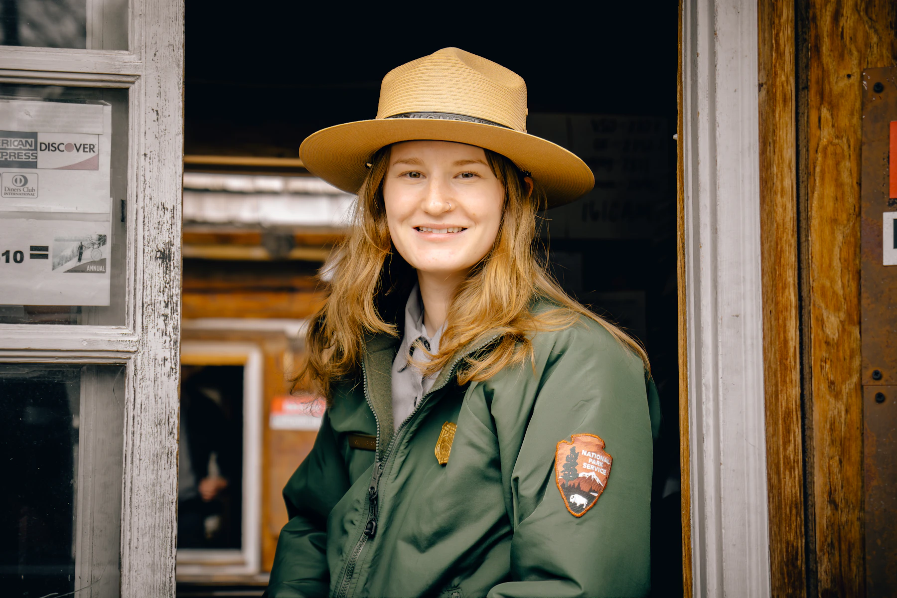 A smiling park ranger in a flat-brimmed hat and green uniform jacket standing in a wooden doorway