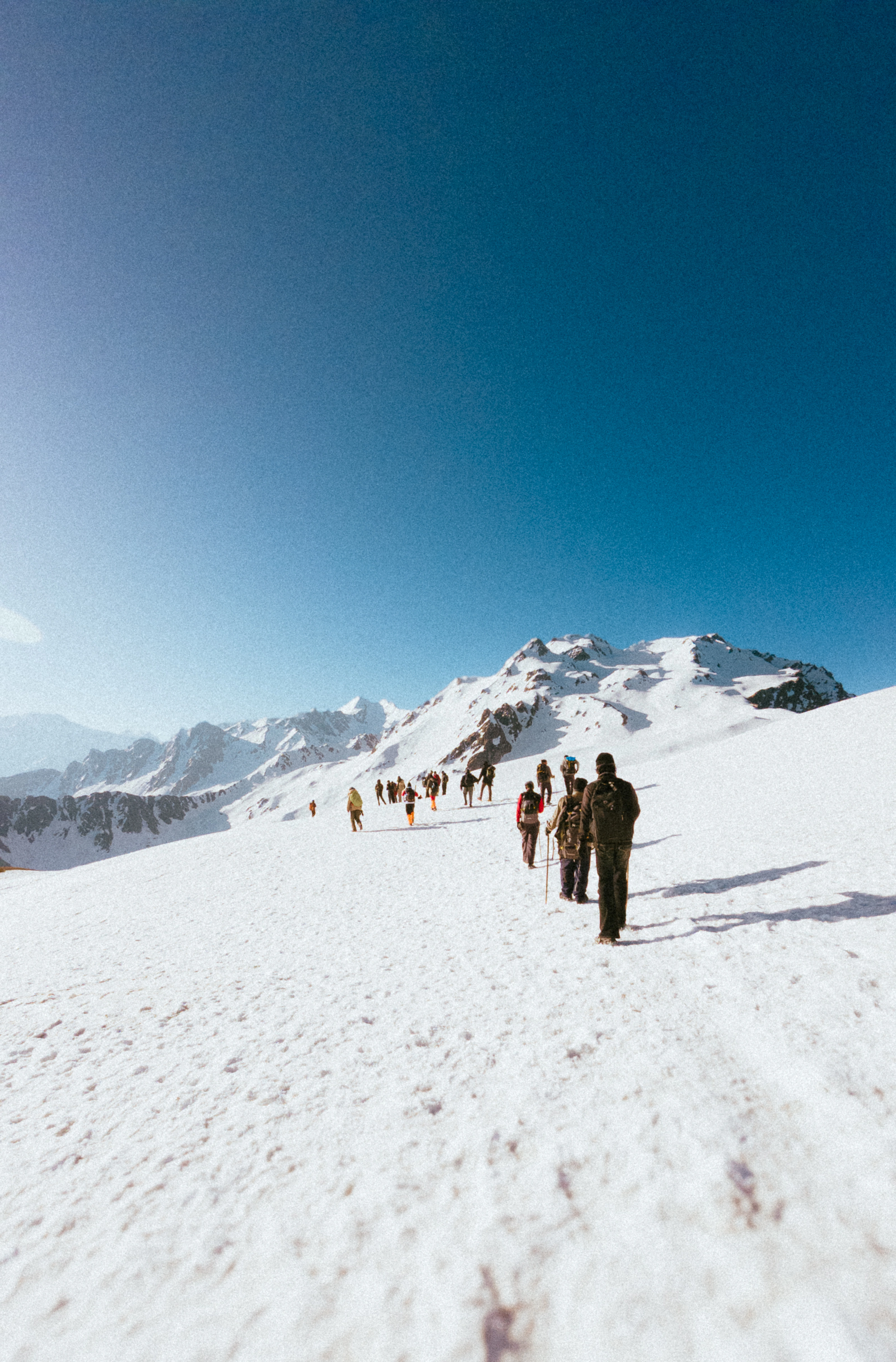 Hikers climbing beneath a treacherous alpine mountain under a clear blue sky, with radiant light above the summit symbolizing hope