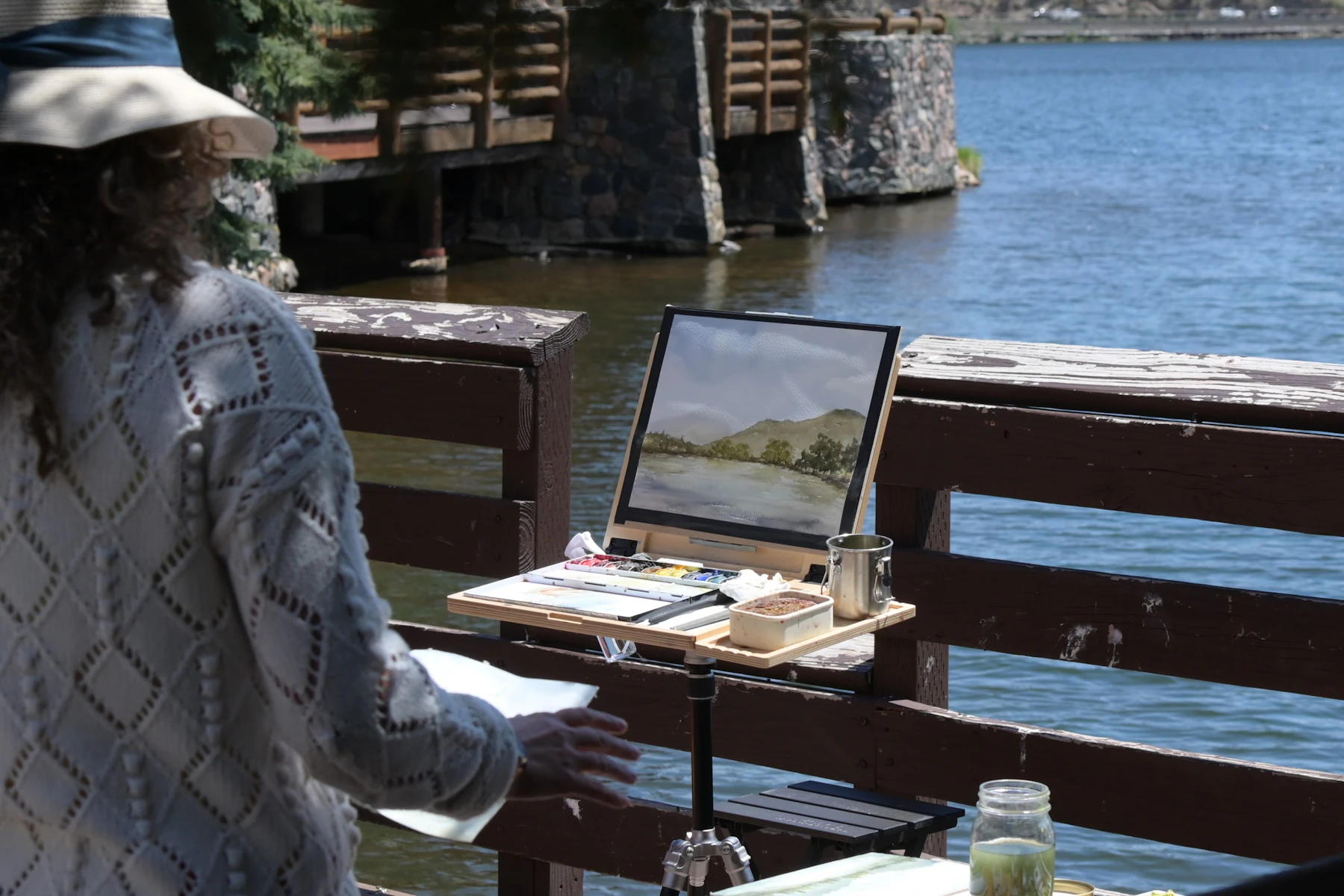 A painter in a sun hat working at a portable easel beside the water