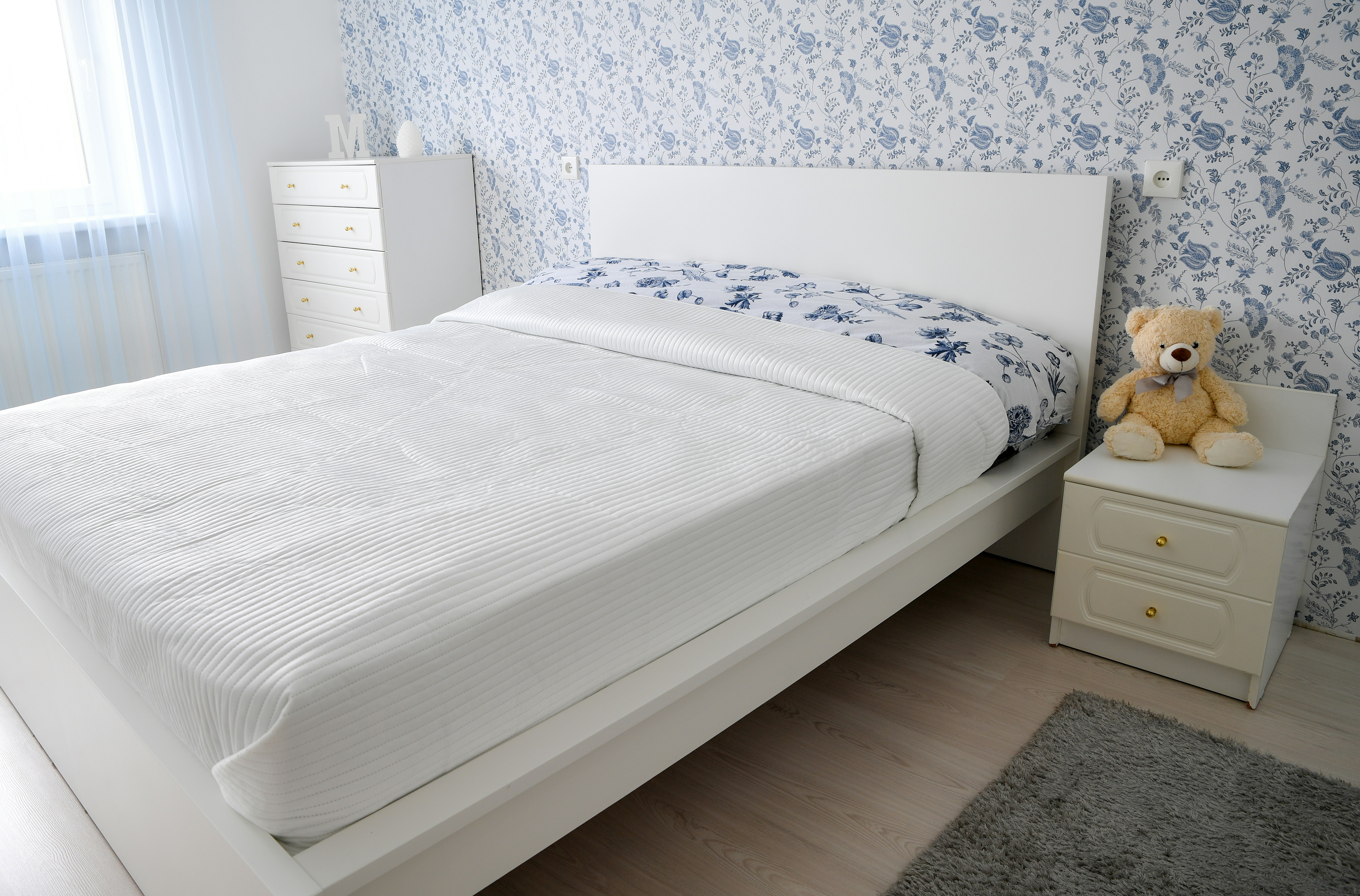 A bright and cozy modern bedroom interior featuring a white double bed, a matching nightstand with a teddy bear, and elegant blue and white floral wallpaper.📸 Photo by me IG: @margoevardson