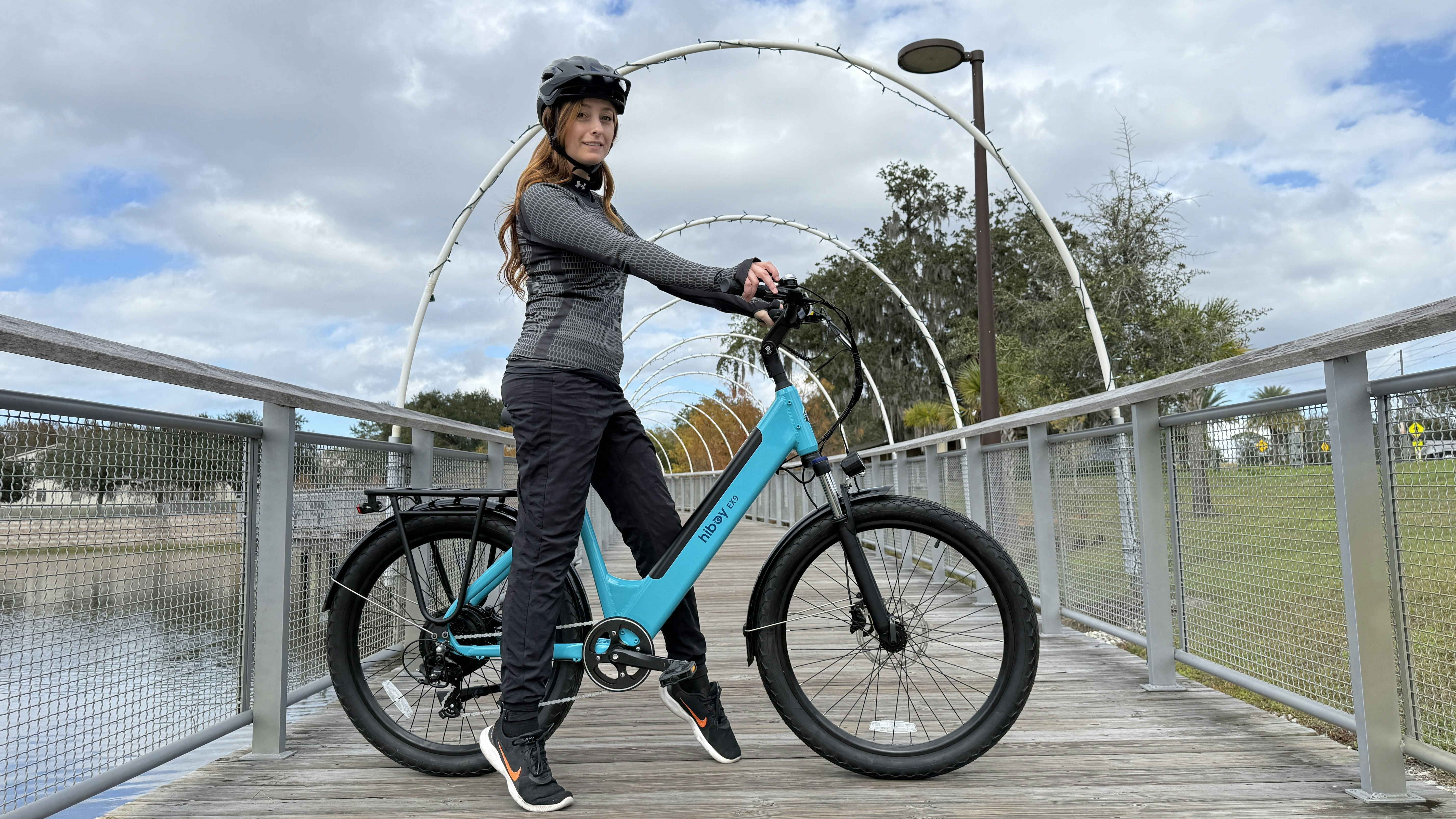 Woman in helmet stands with teal bicycle on bridge
