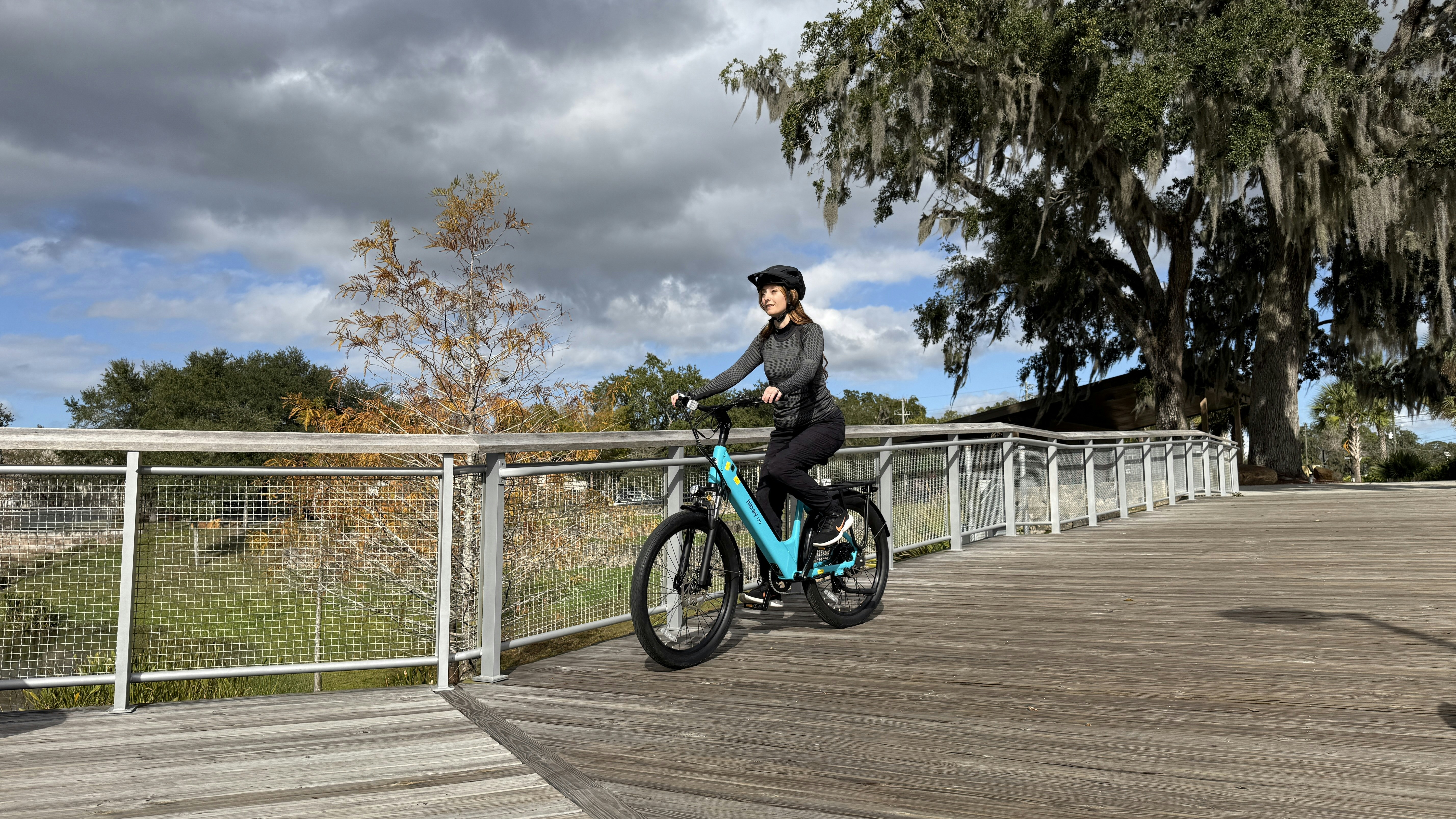 A woman rides a teal bicycle on a wooden boardwalk