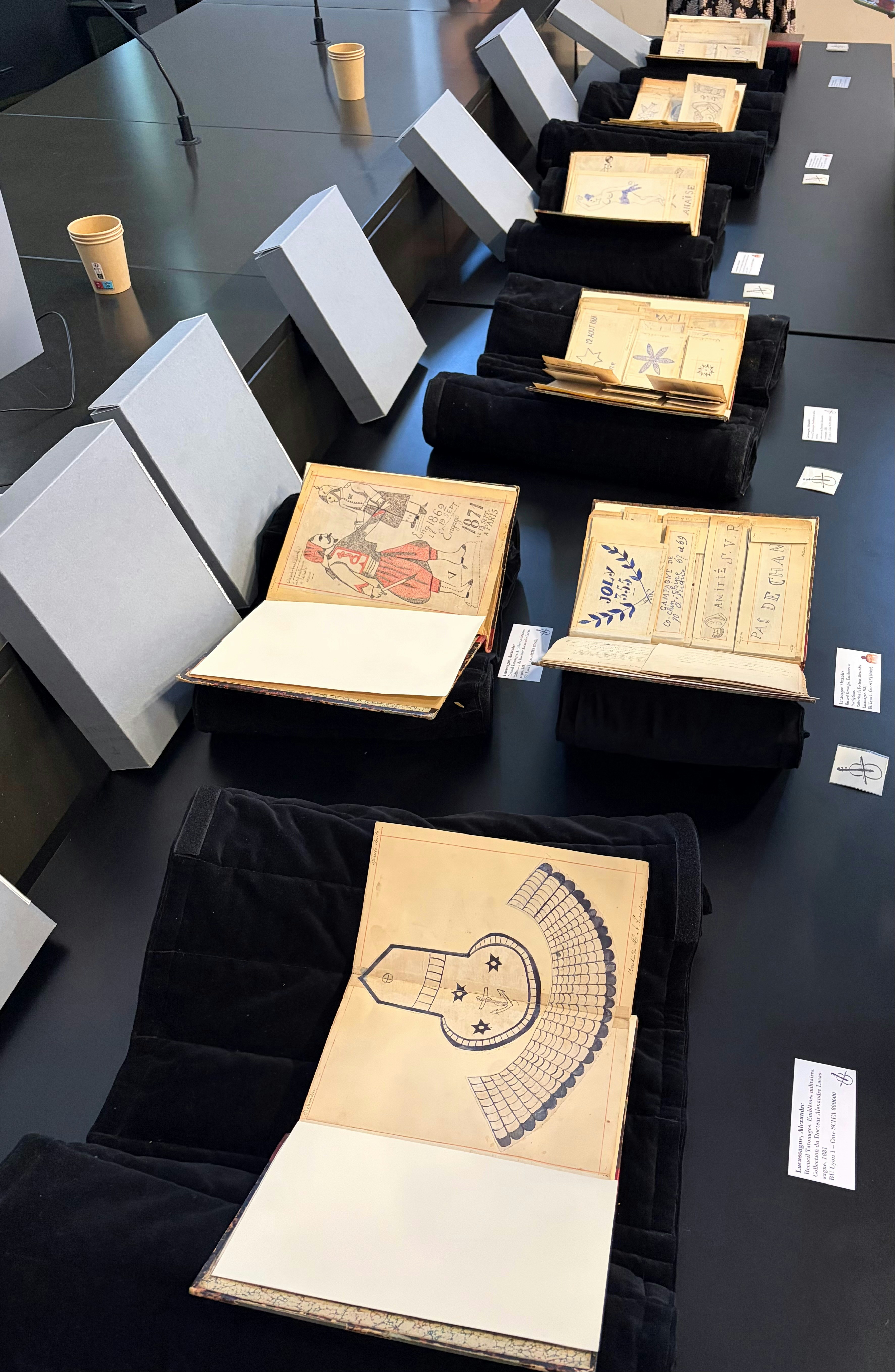 Plusieurs carnets de tatouages d'Alexandre Lacassagne ouverts et présentés sur une table lors de la présentation à l'université