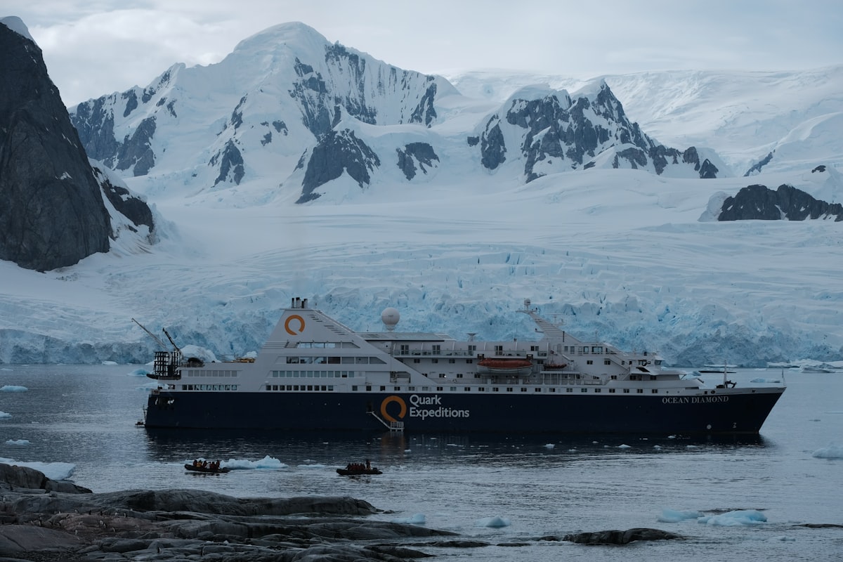 A dark-hulled expedition ship lying at anchor in front of a wide glacier face, with two small inflatable boats in the water beside it and snow-covered peaks rising behind