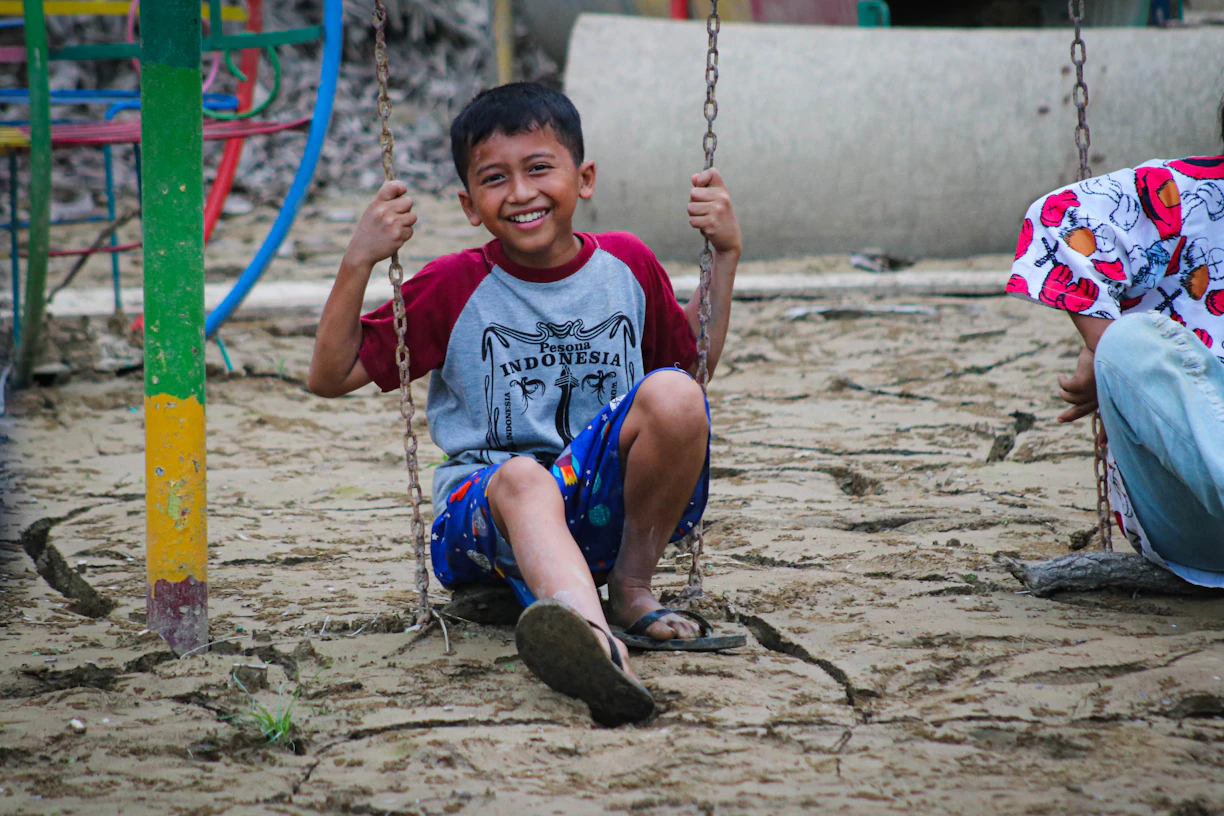 Happy boy smiles on a swing in a sandy playground.