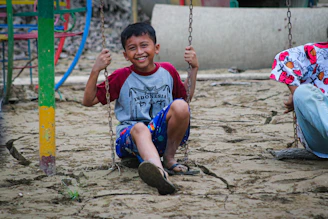 Happy boy smiles on a swing in a sandy playground.
