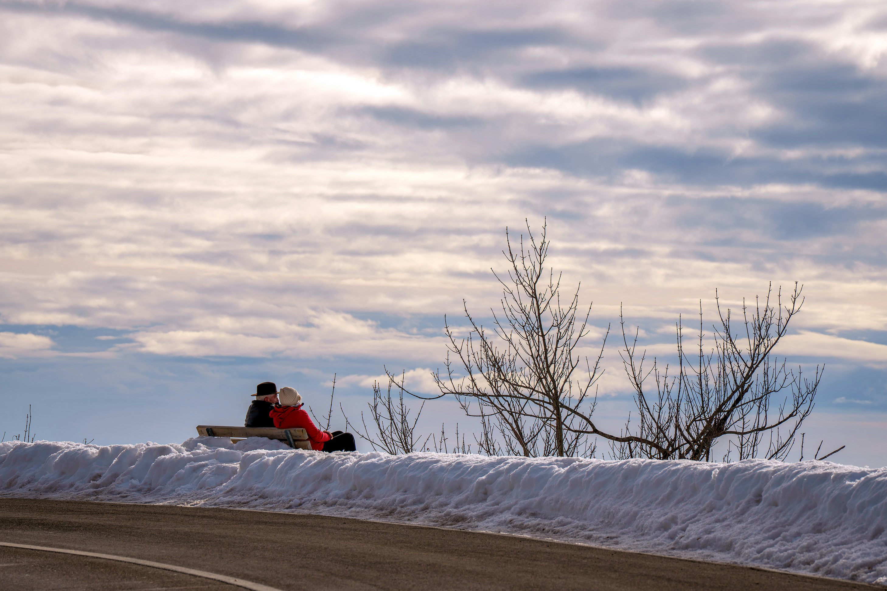 Two people sit on a bench by a snowy road.