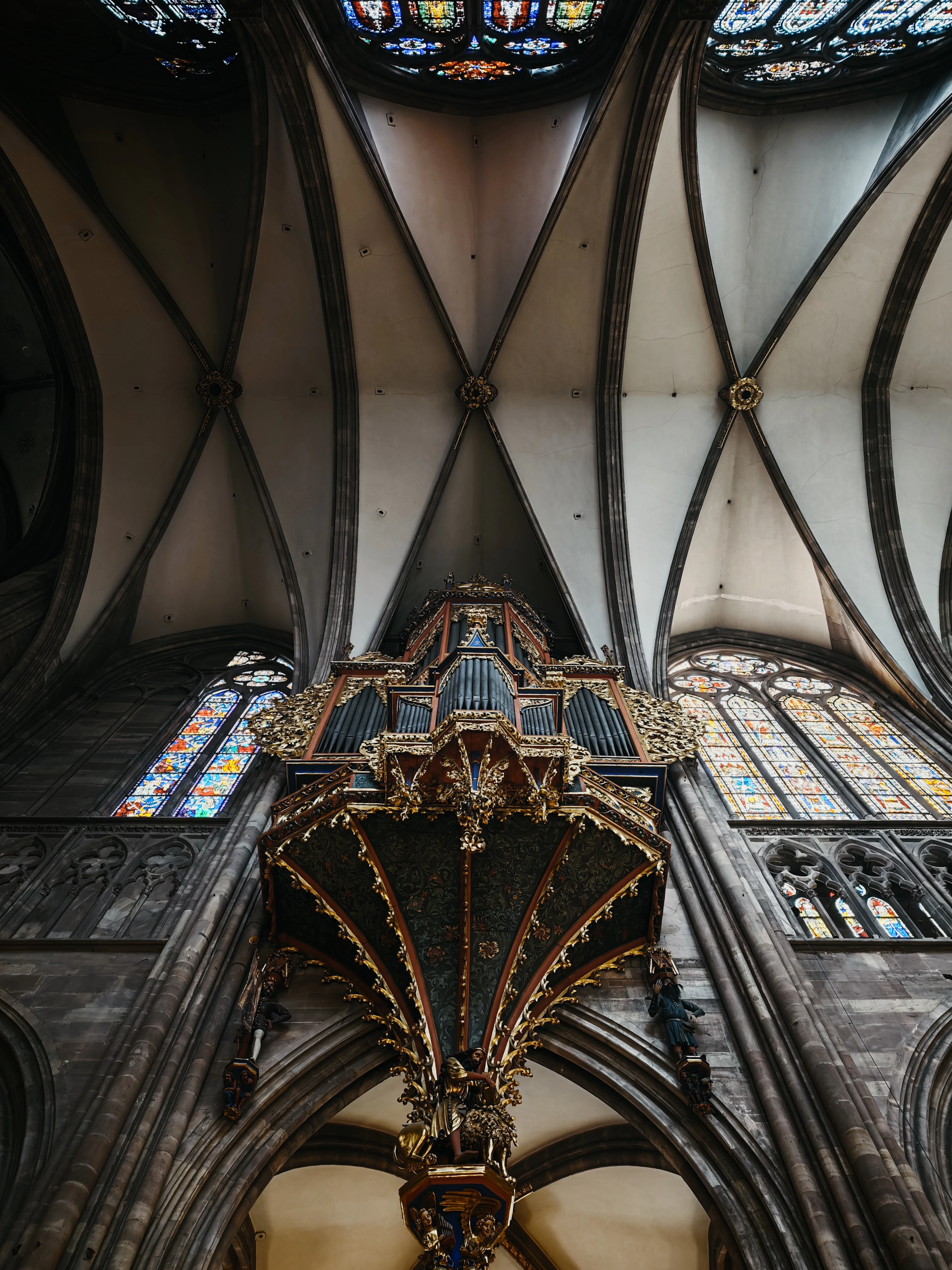 Looking straight up inside Strasbourg Cathedral at the gilded swallow's-nest organ hanging from the nave wall, ribbed vaults and stained glass spreading above it.