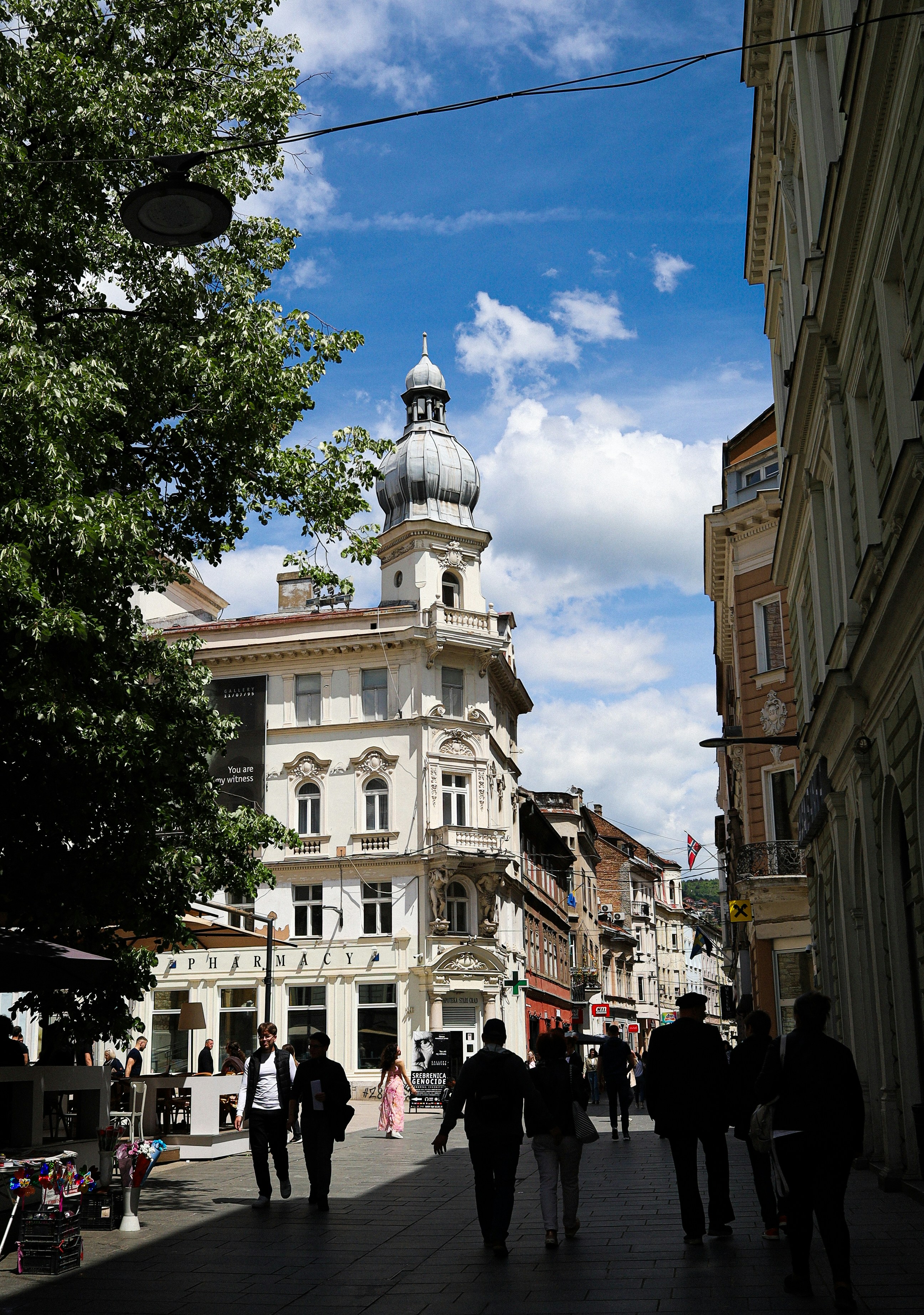 Pedestrians stroll past ornate buildings on a sunny european street.