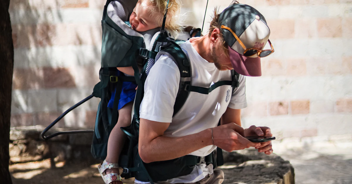 Parent carrying a toddler in a hiking baby carrier outdoors on a sunny day.