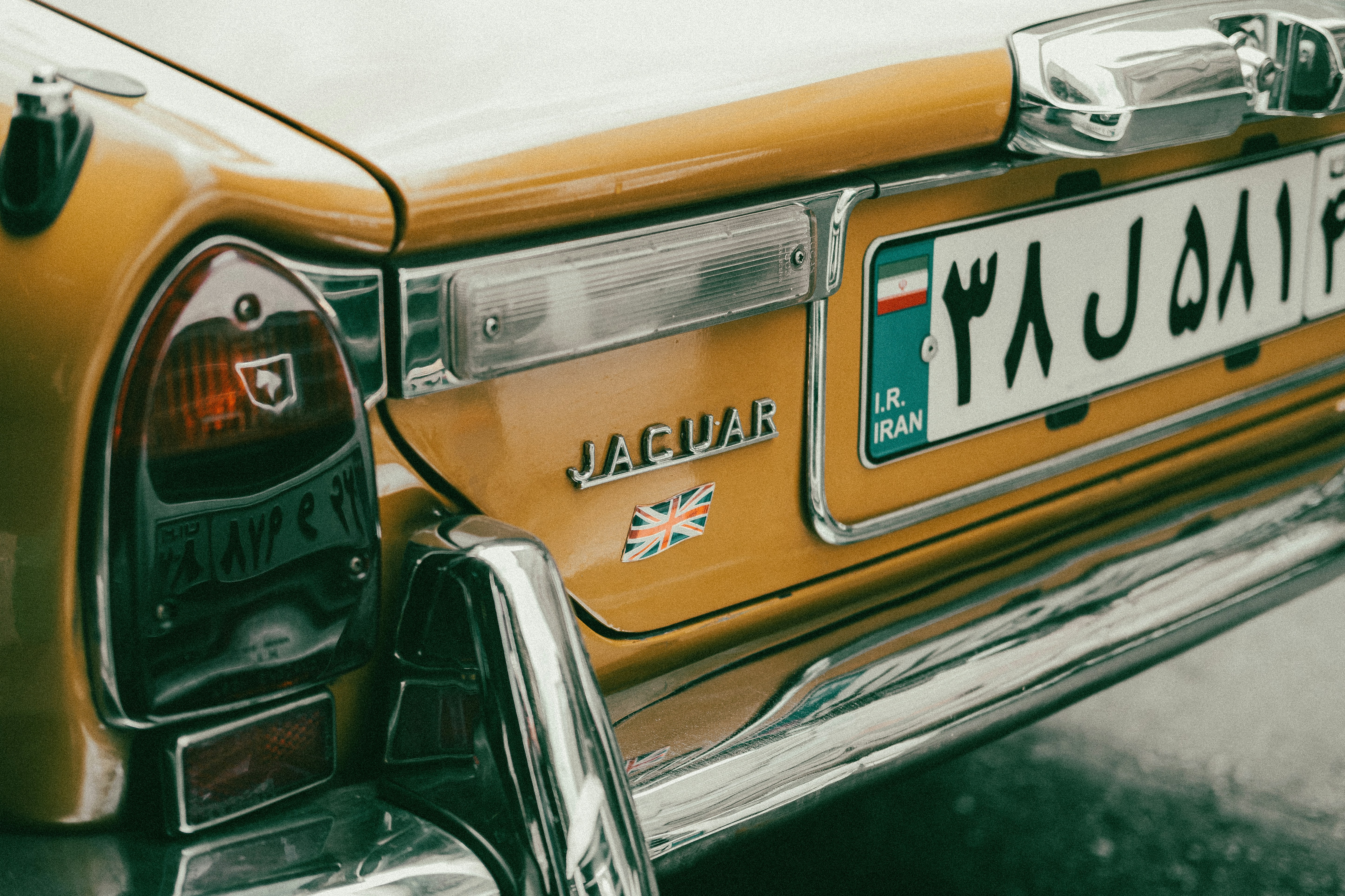 Close-up of a vintage orange Jaguar rear with chrome bumper, emblem, and Iranian license plate; the focus is on branding badge and tail light details, capturing color and chrome reflections in a classic automotive setting.