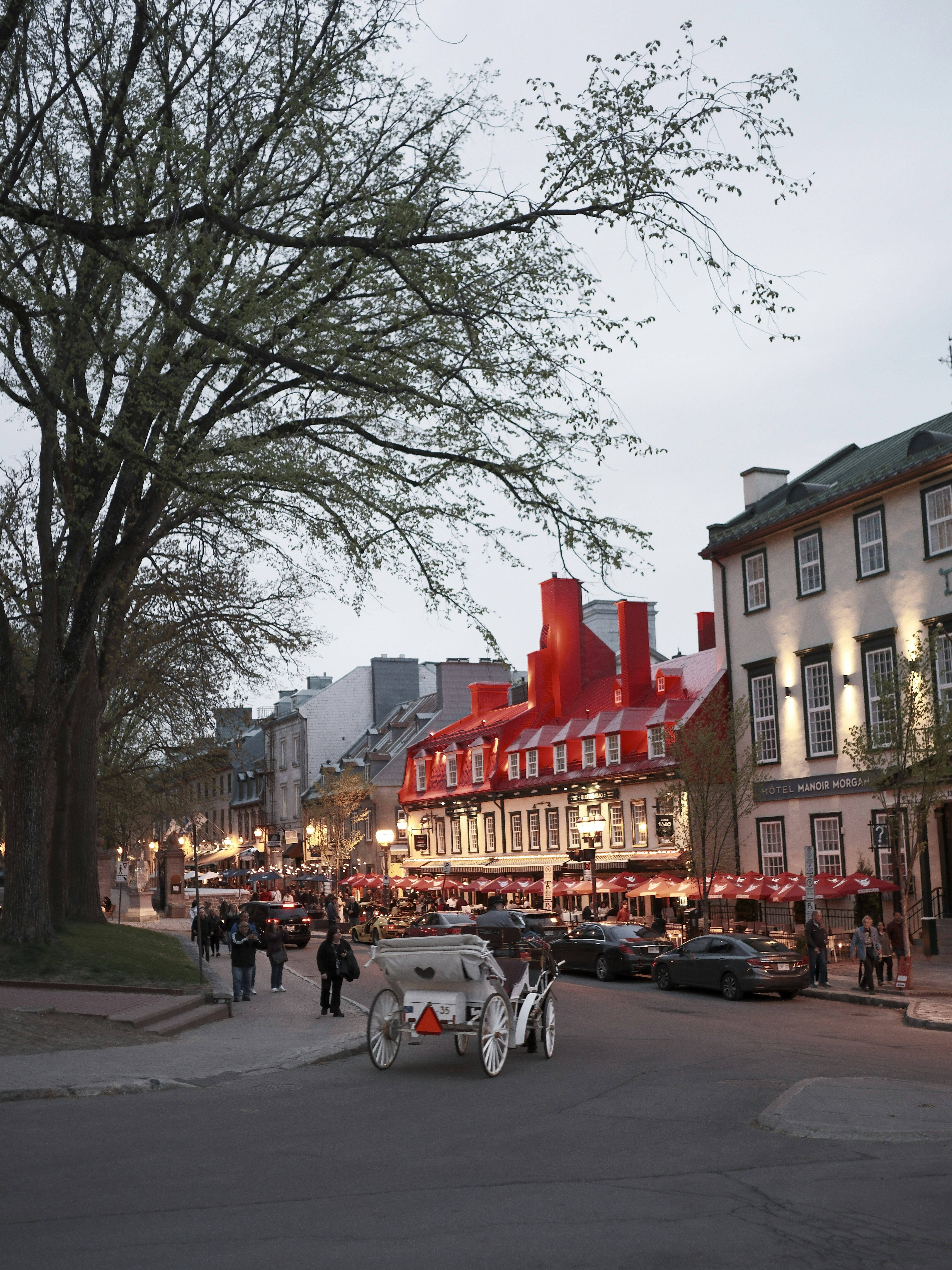 Historic street with red-roofed façade buildings and a horse-drawn carriage in the foreground; pedestrians and cars line the lively avenue at dusk.