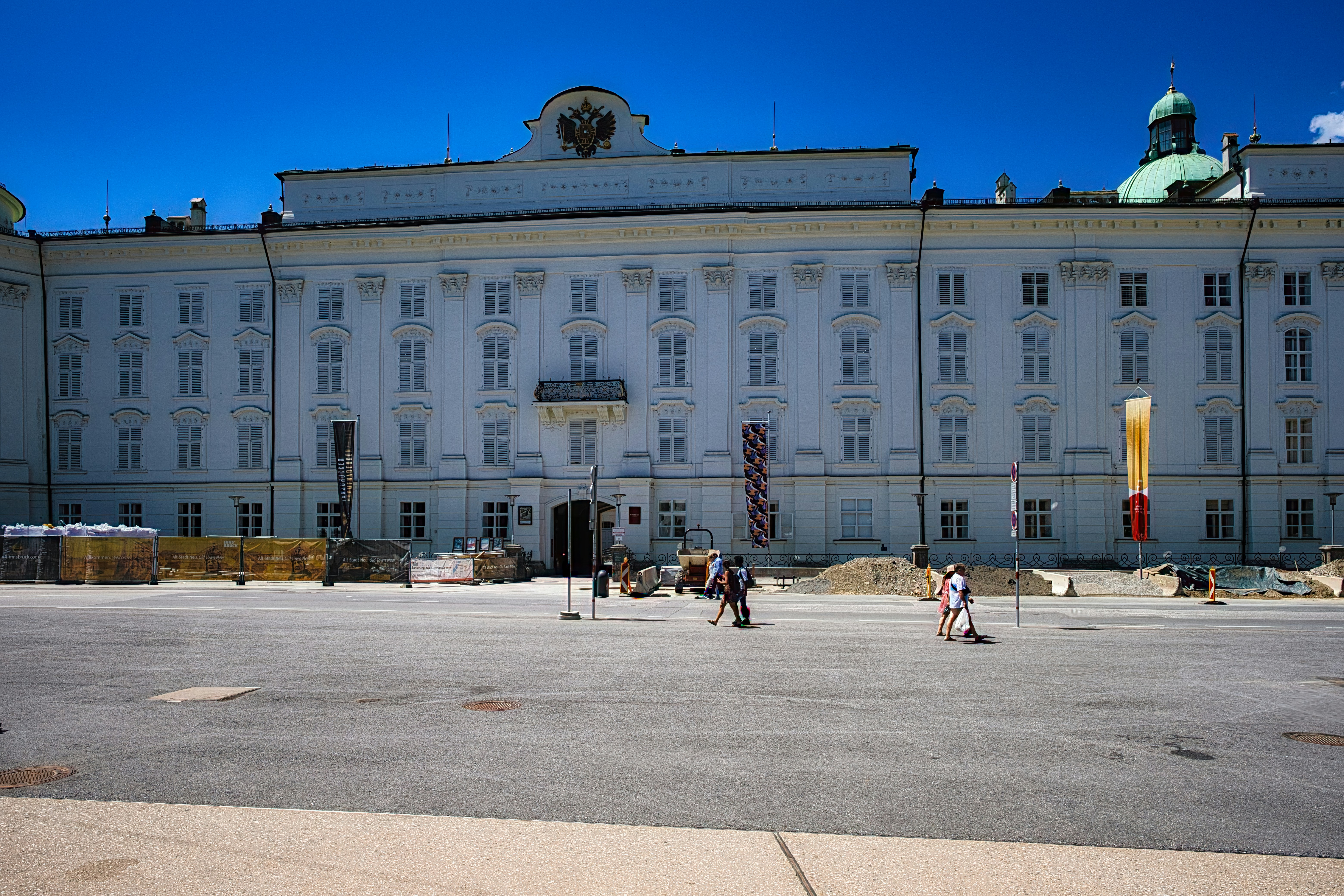 Wide-angle view of a grand white-baroque building with a central pediment,-set under a bright blue sky; pedestrians and street furniture line a broad plaza in front, indicating a public square under renovation.