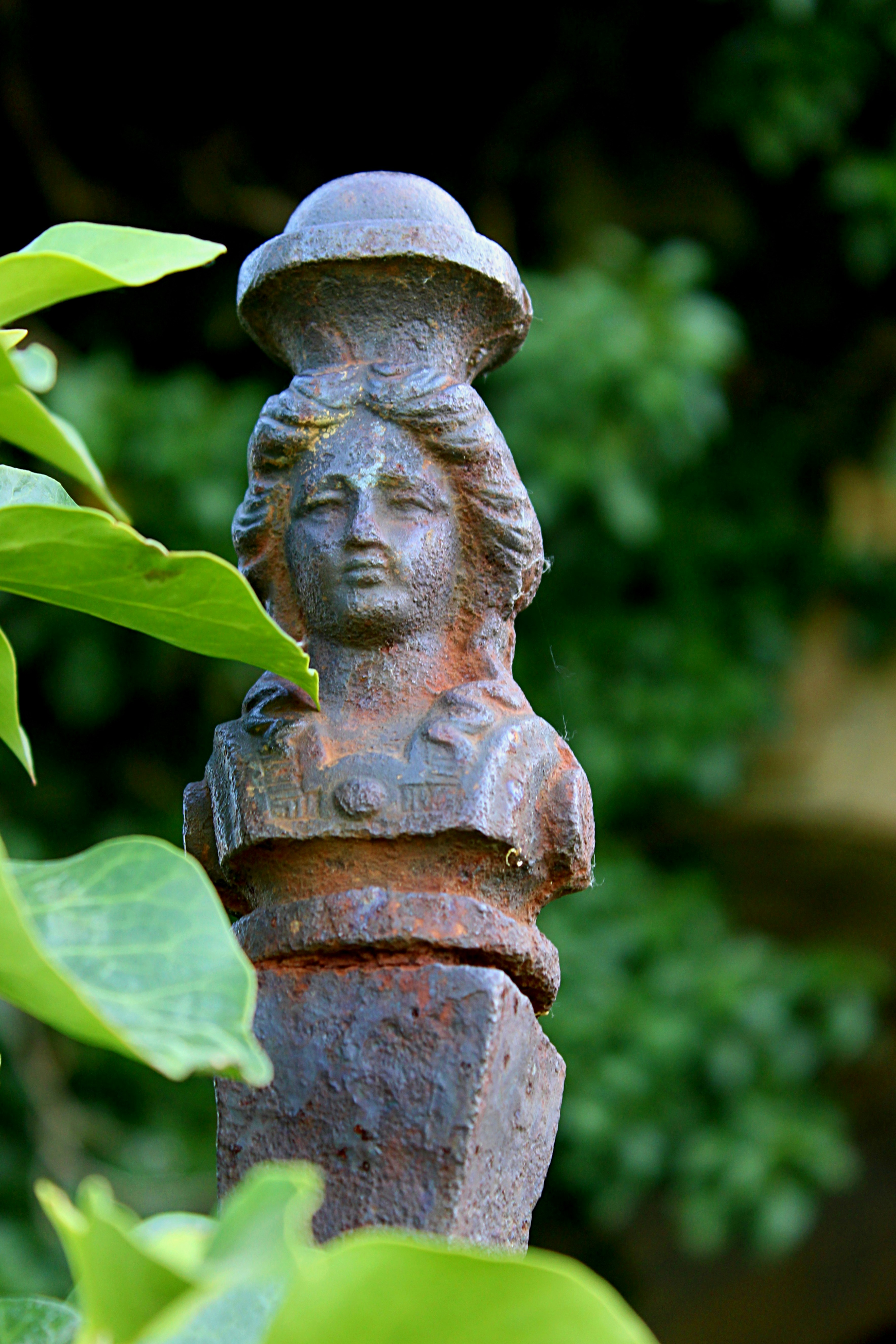 Rusty metal bust of a woman peeking through green leaves.