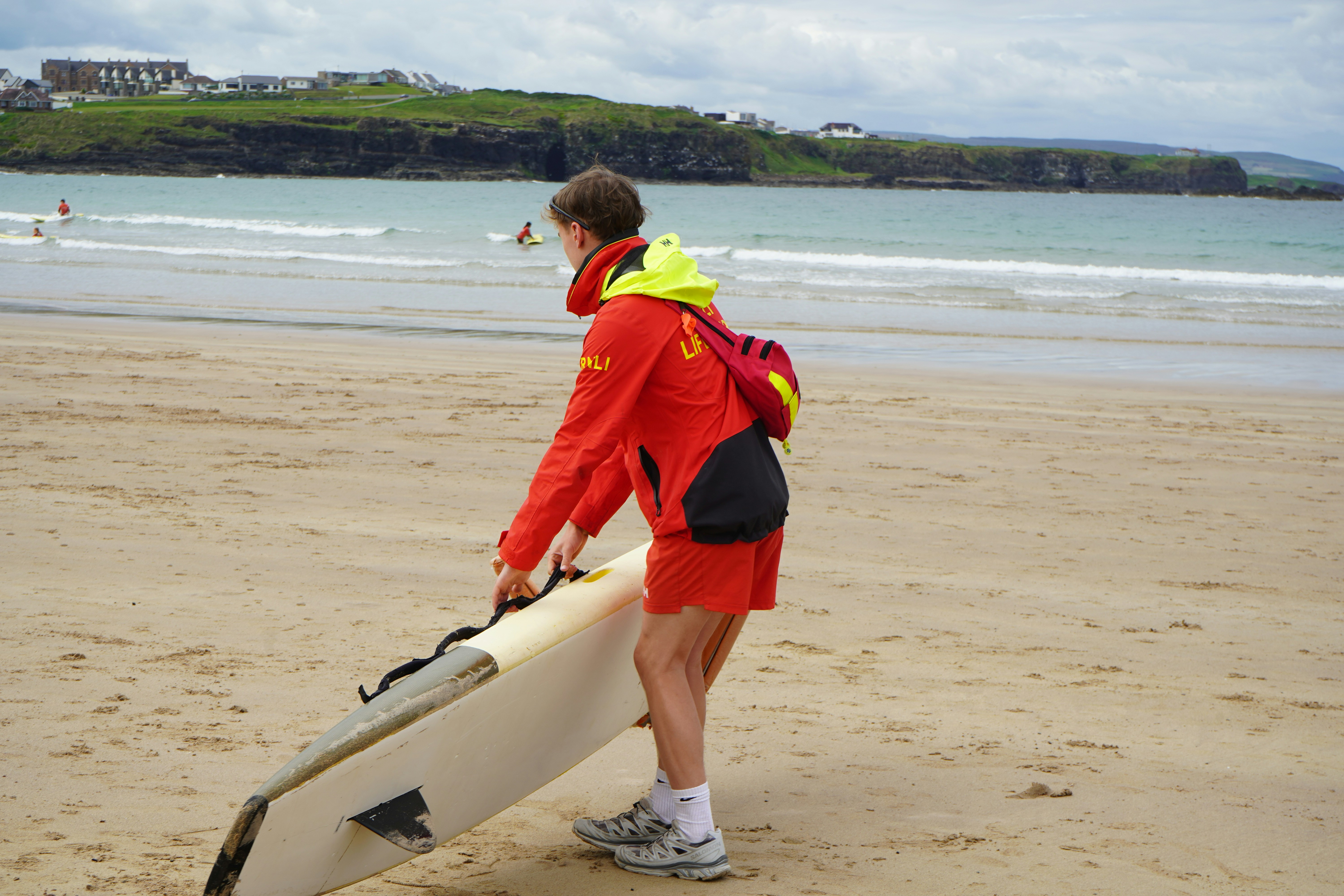 Lifeguard carrying a surfboard on a sandy beach.