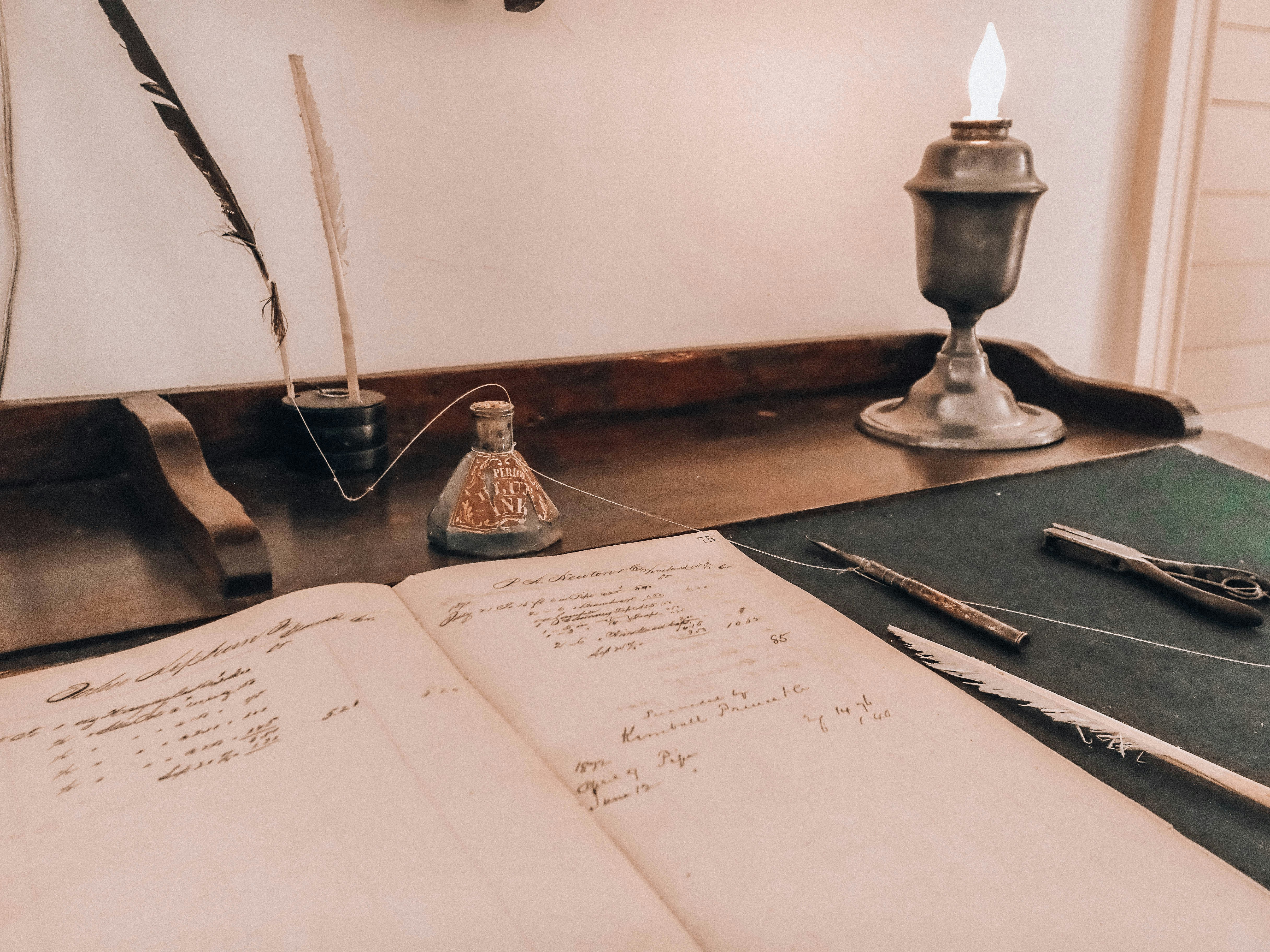 Vintage desk with open book, quill, inkwell, and lit lamp.