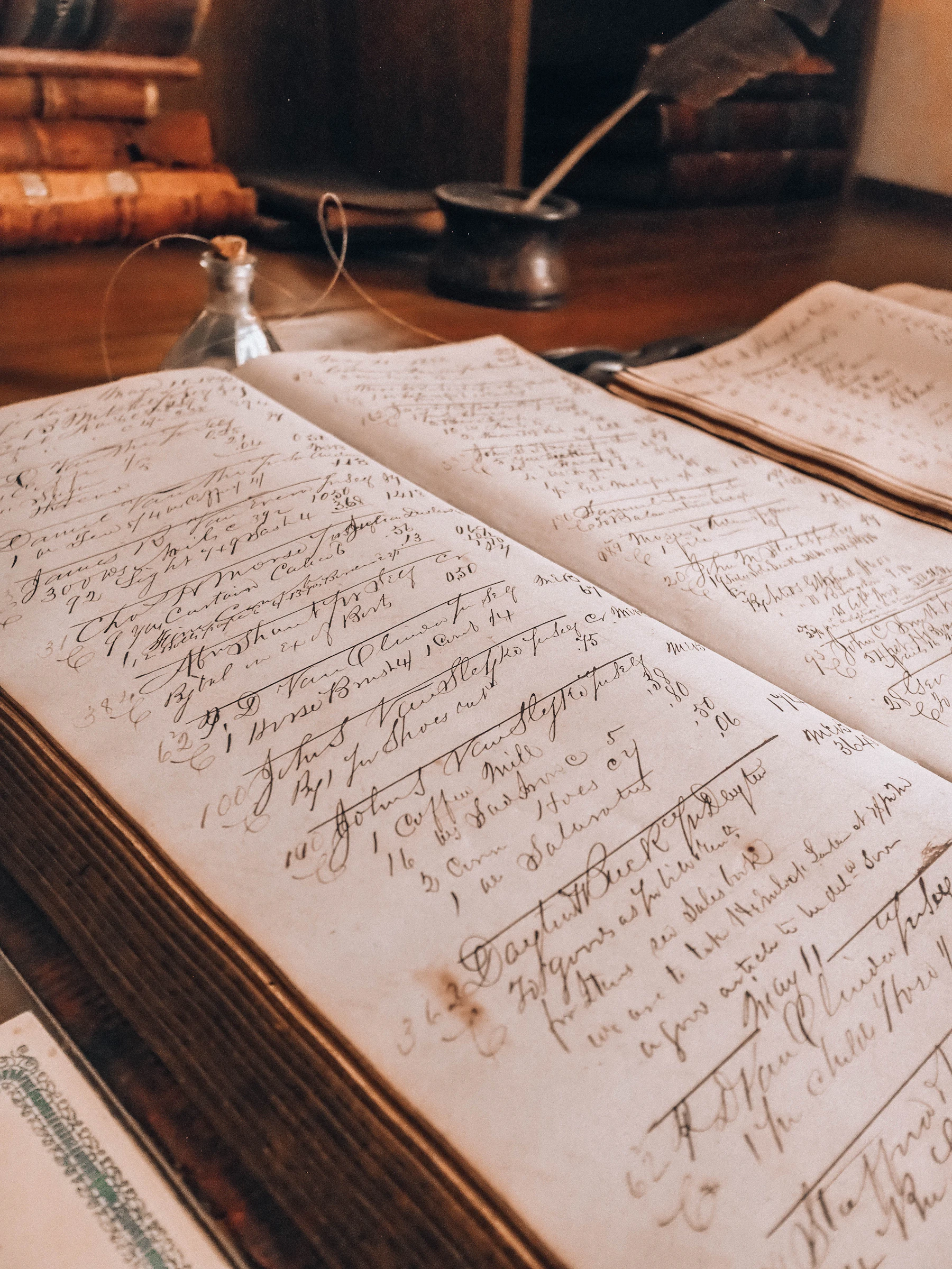 An open ledger of handwritten entries on a wooden desk, an inkwell and a quill behind it