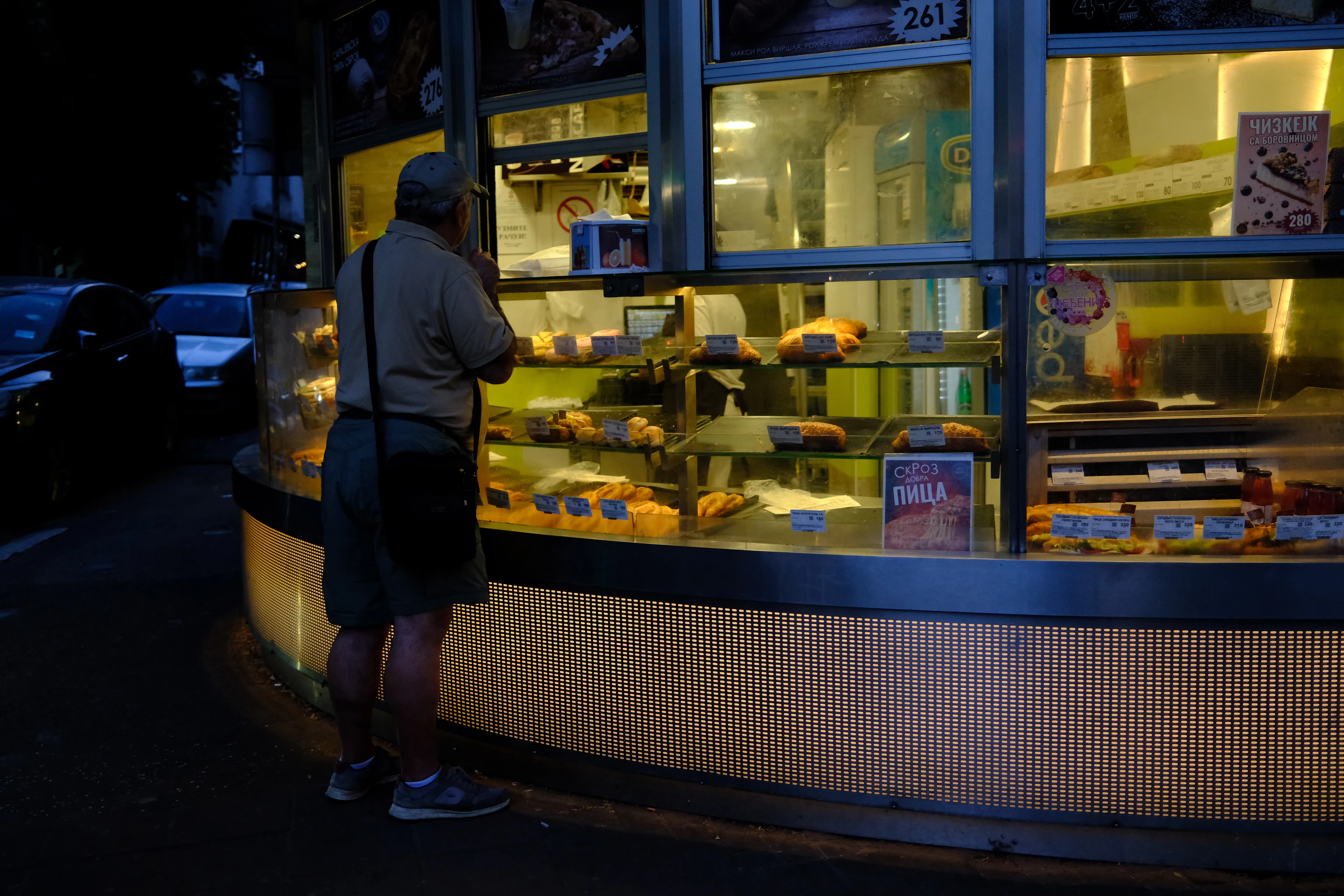 A man leans in to study the trays of pastries in the lit glass counter of a late-night Belgrade bakery kiosk, the warm yellow interior glowing against the dark street behind him.