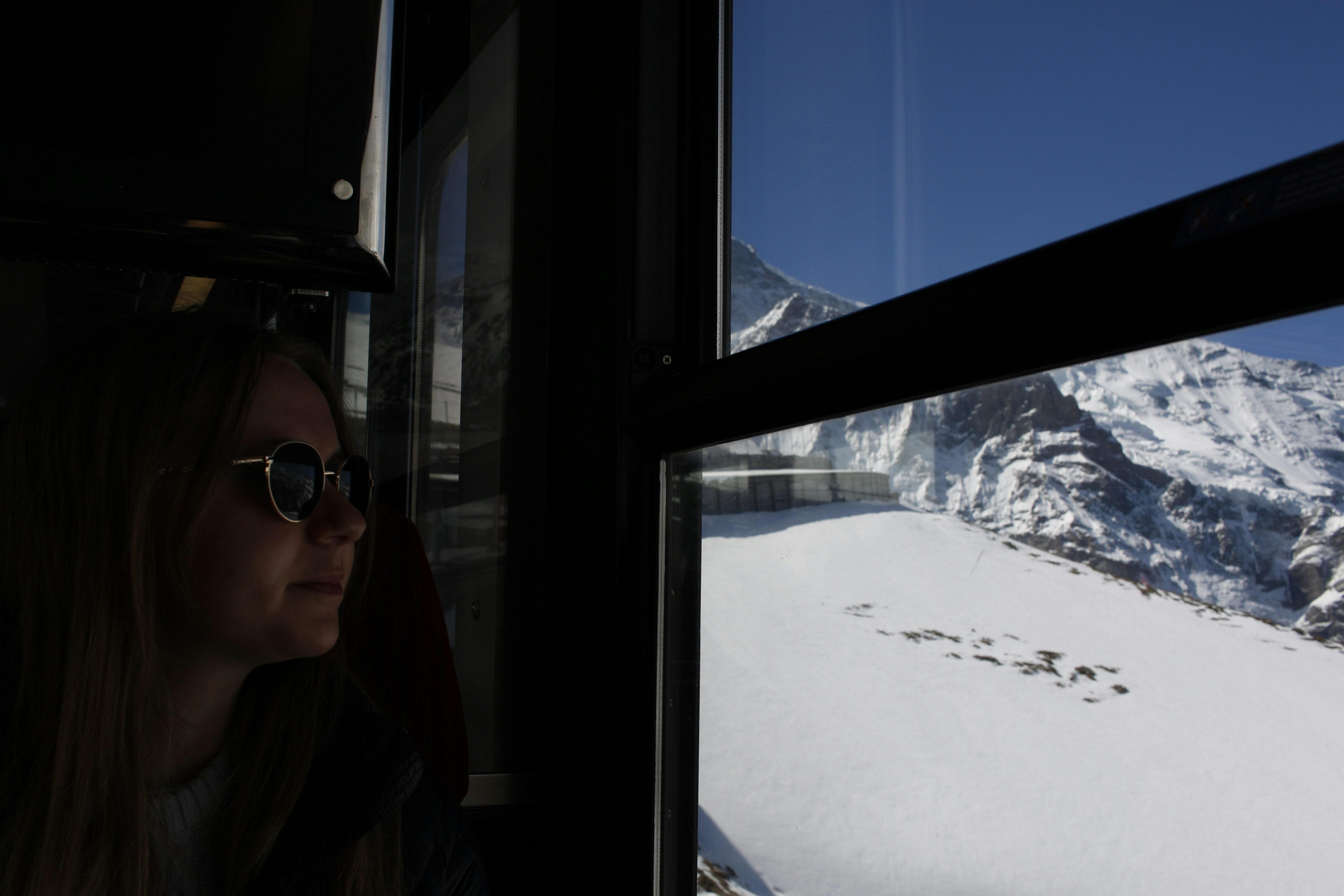 Frau mit Brille blickt aus dem Zugfenster auf verschneite Hänge und Berggipfel unter blauem Himmel