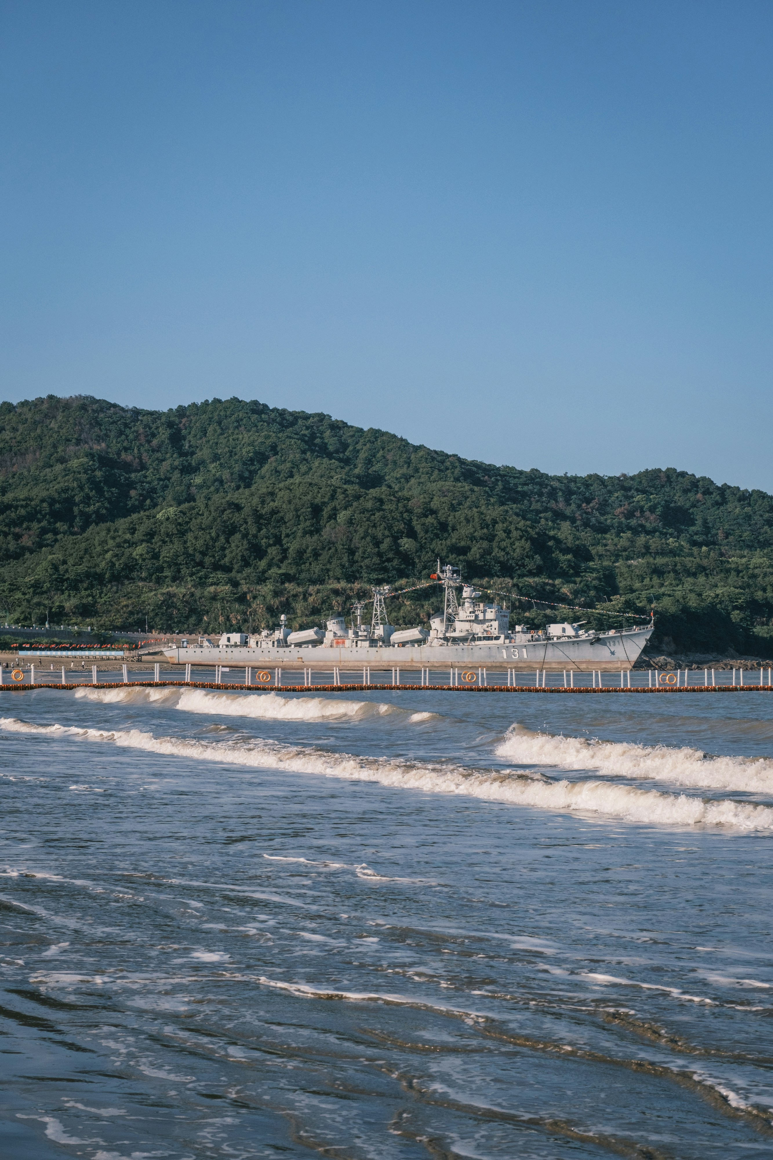 Warship by a beach with waves and a forested mountain.