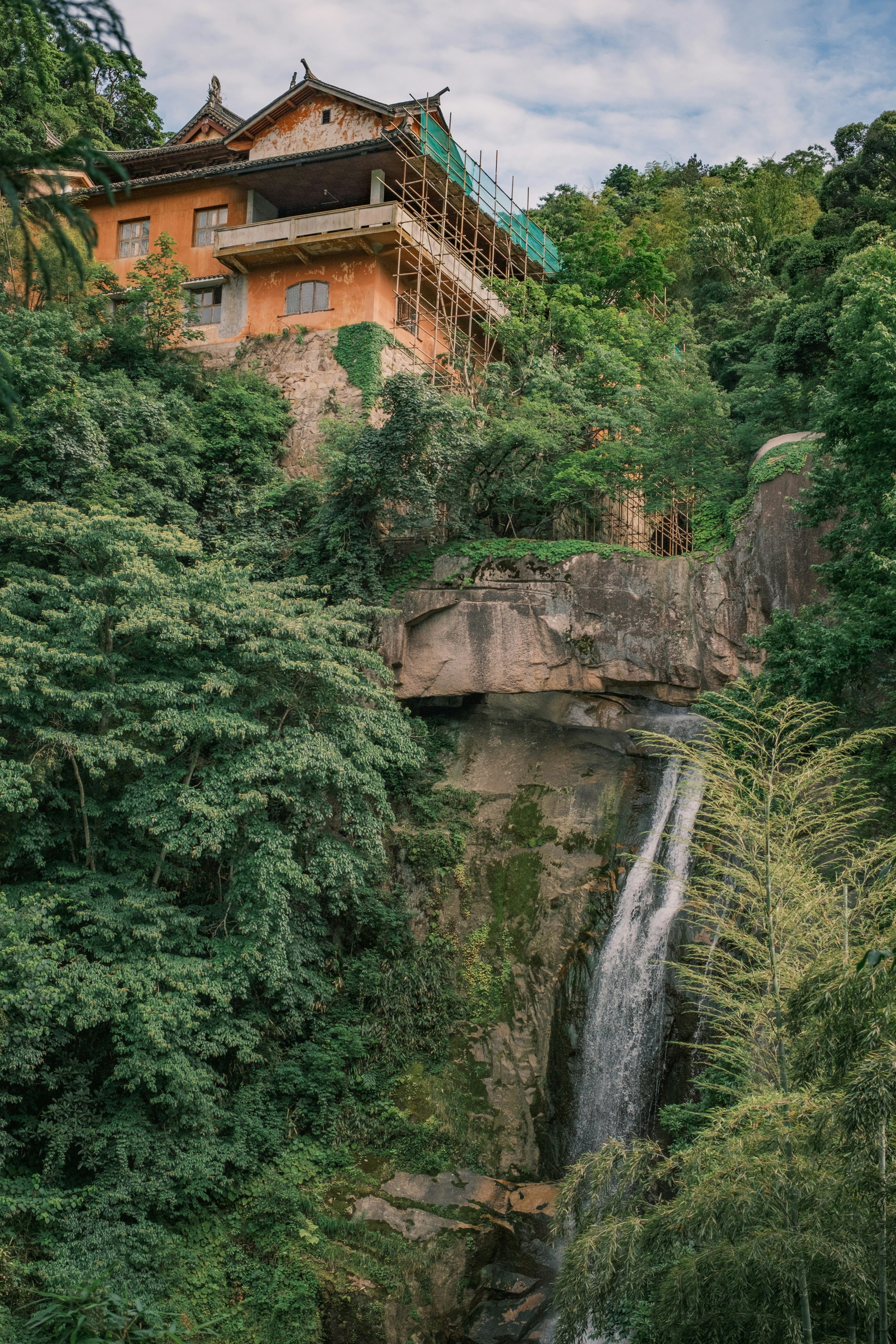 Traditional building perched above a waterfall in lush green mountains.