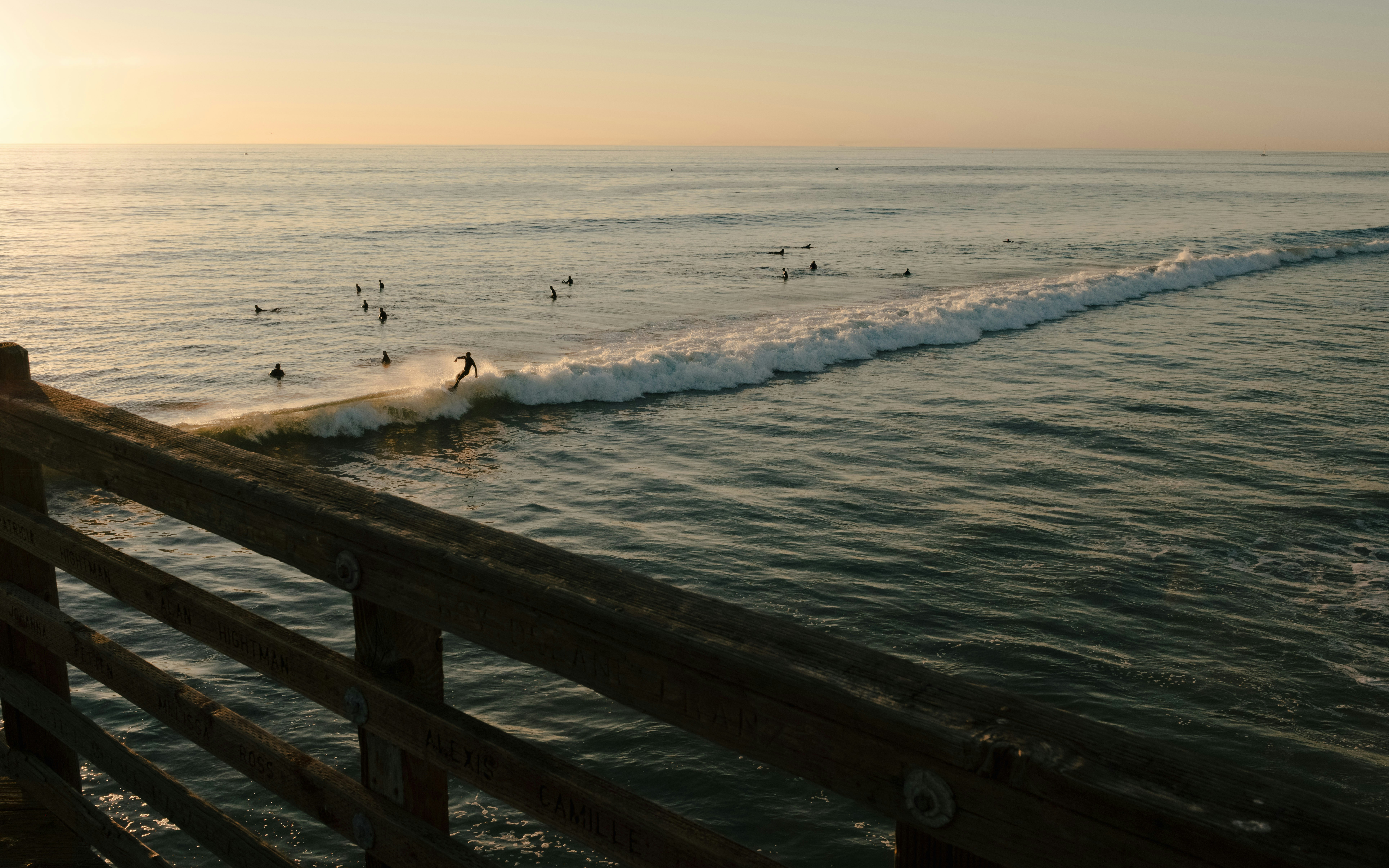 Surfers ride waves near a pier at sunset.