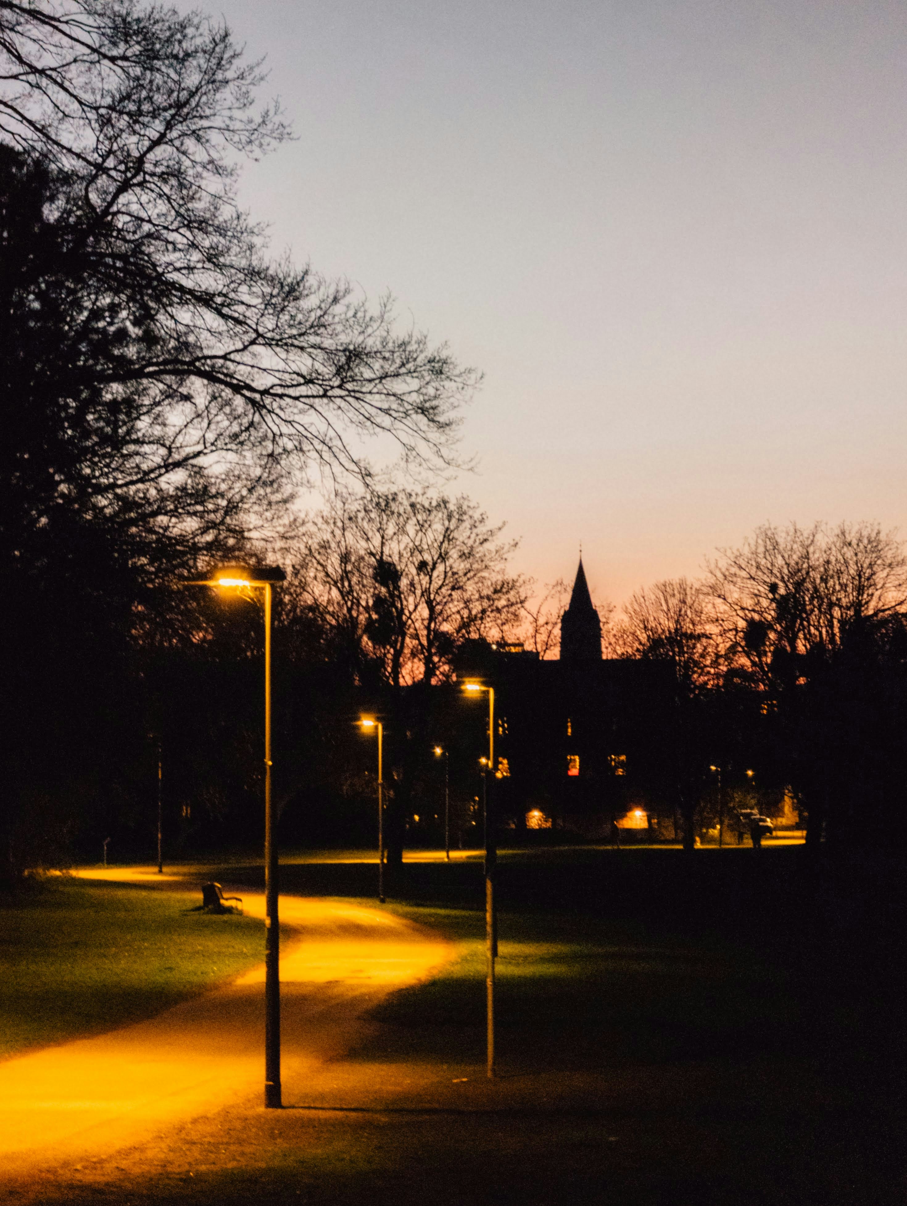 Erleuchteter Parkweg bei Einbruch der Dämmerung mit einer entfernten Gebäudesilhouette.