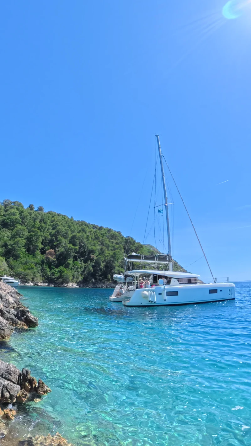 A white sailing catamaran at anchor in a turquoise cove below green hills.