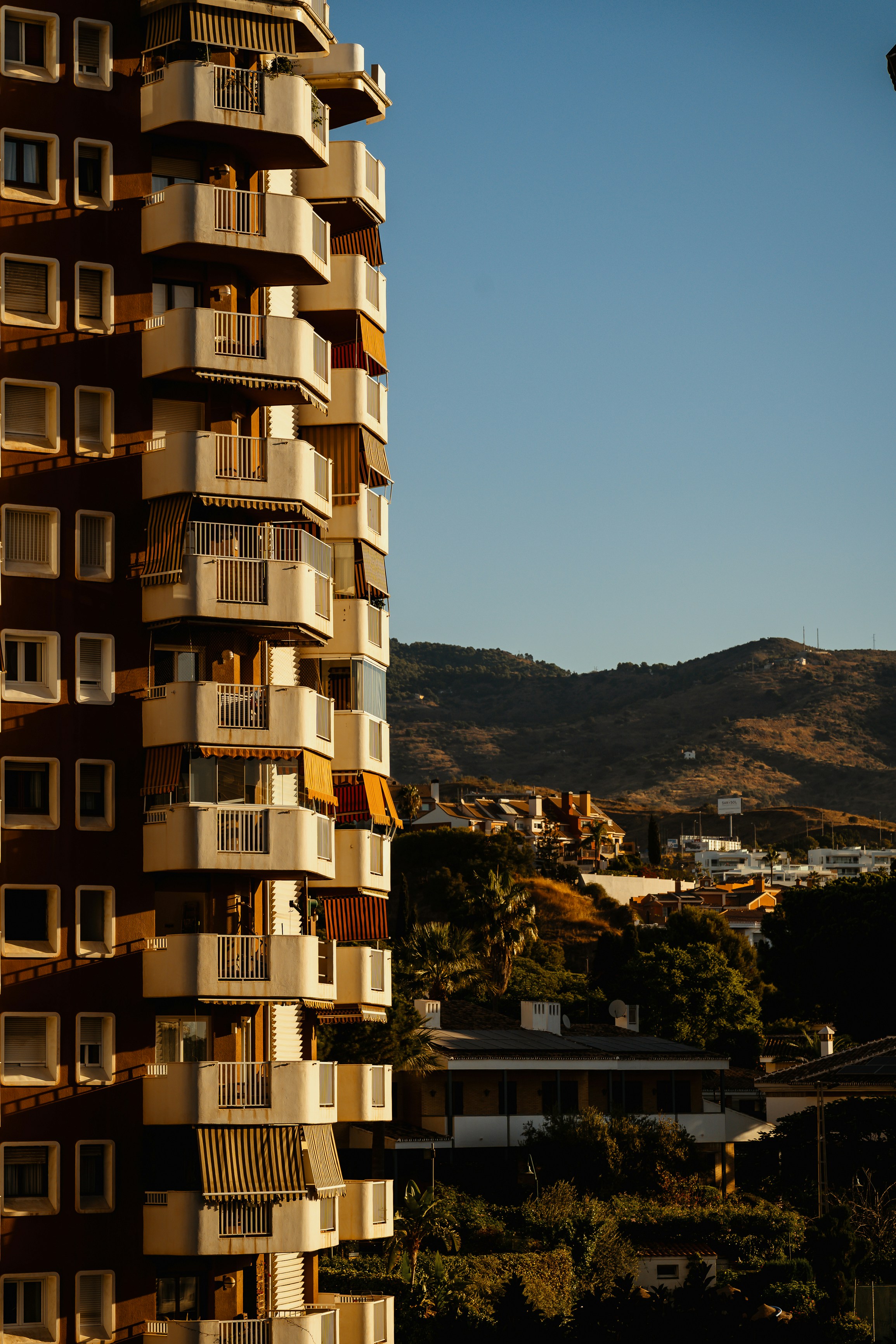 Tall building with balconies, clear sky, and distant hills.