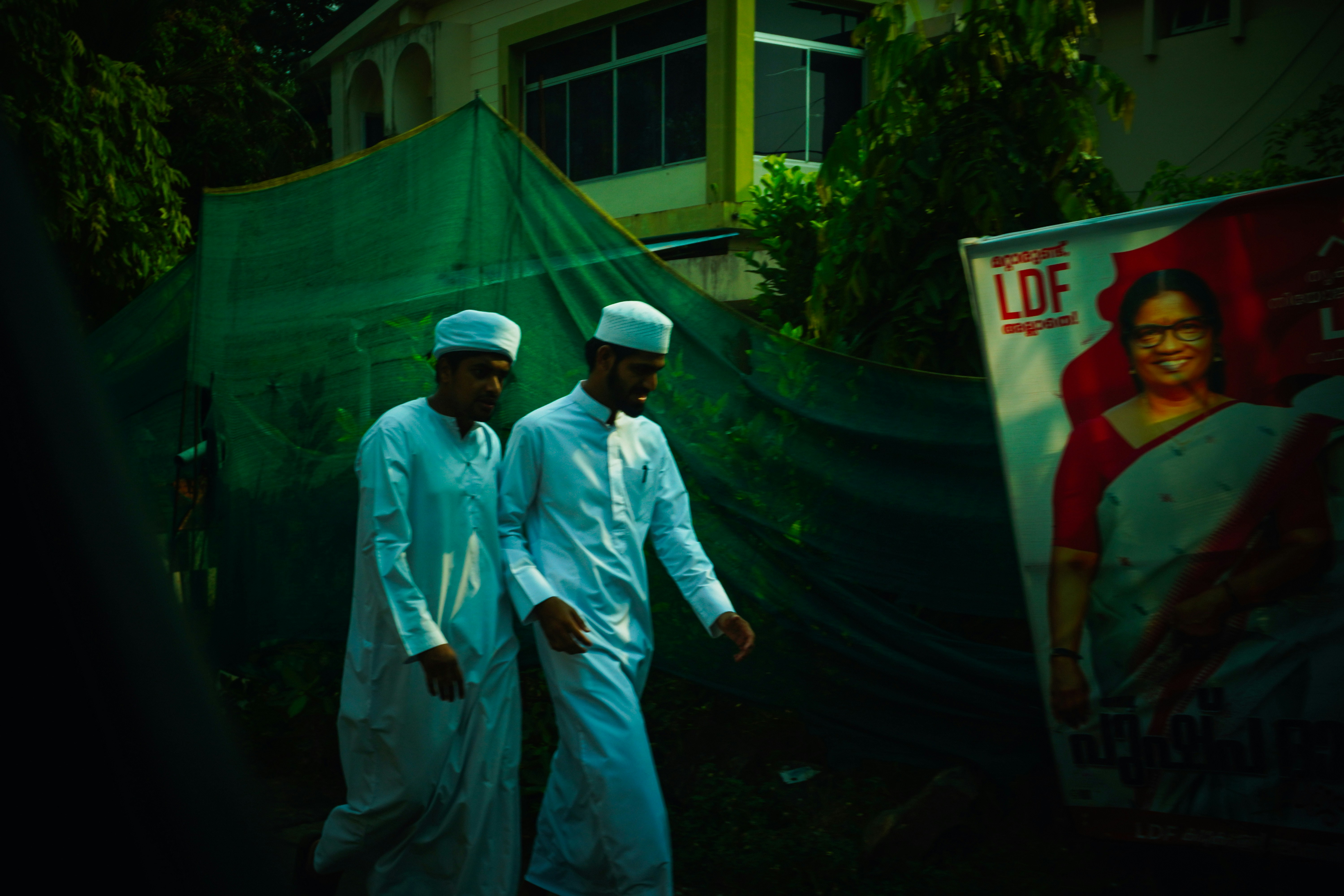 Men in white robes walk past a large political advertisement.