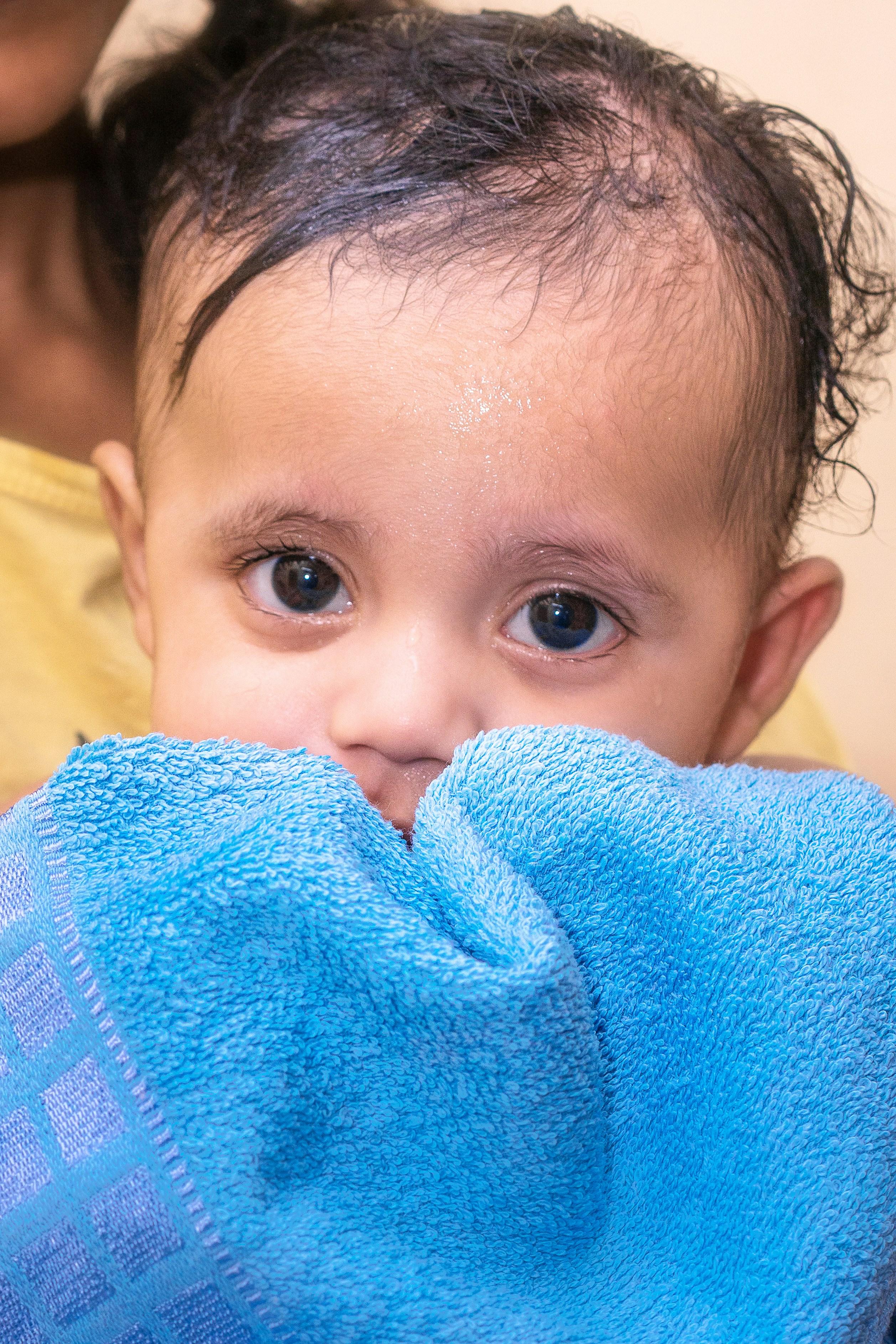 Adorable baby with dark eyes peeking from behind blue towel.