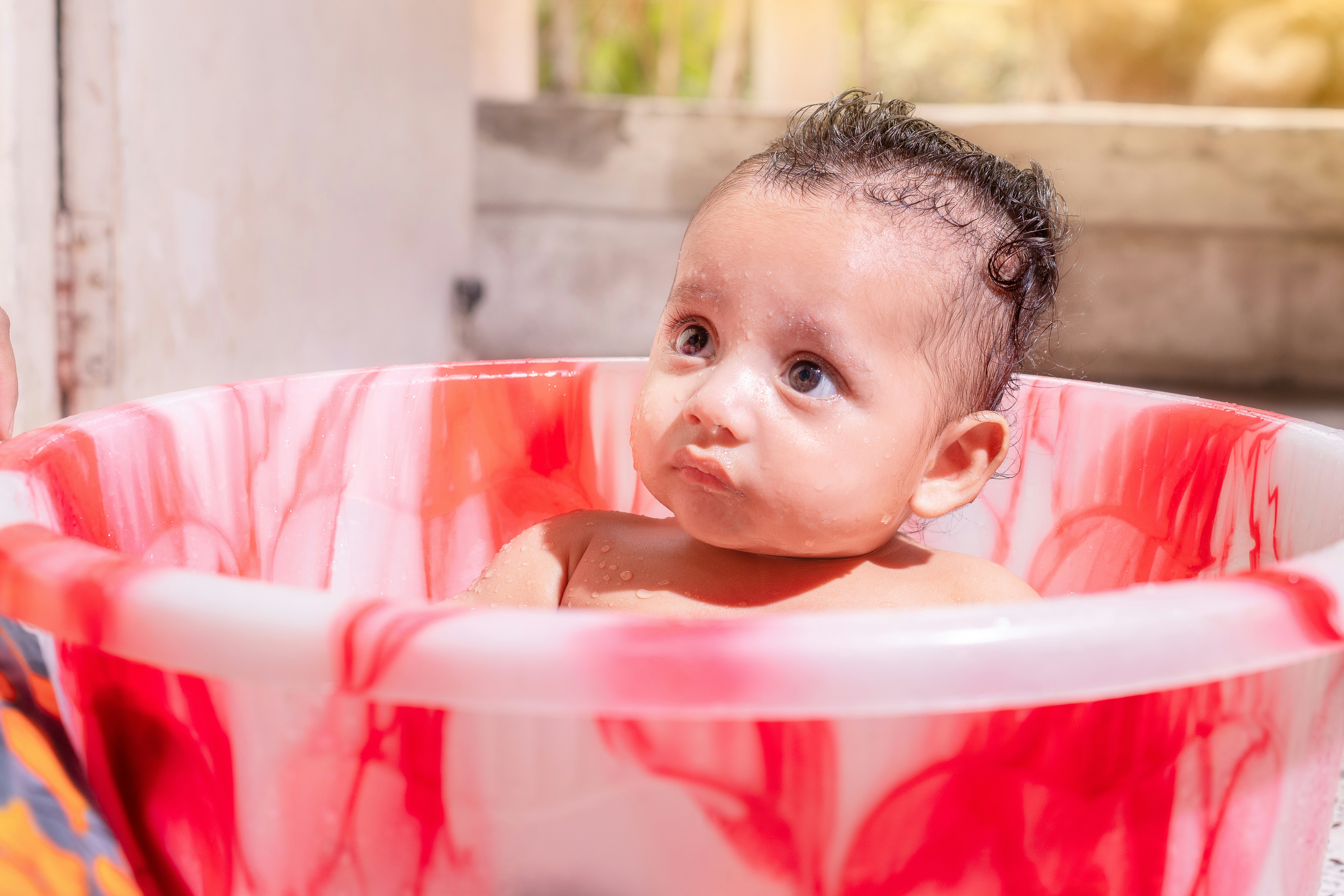 Adorable baby sitting in a red patterned bath tub.