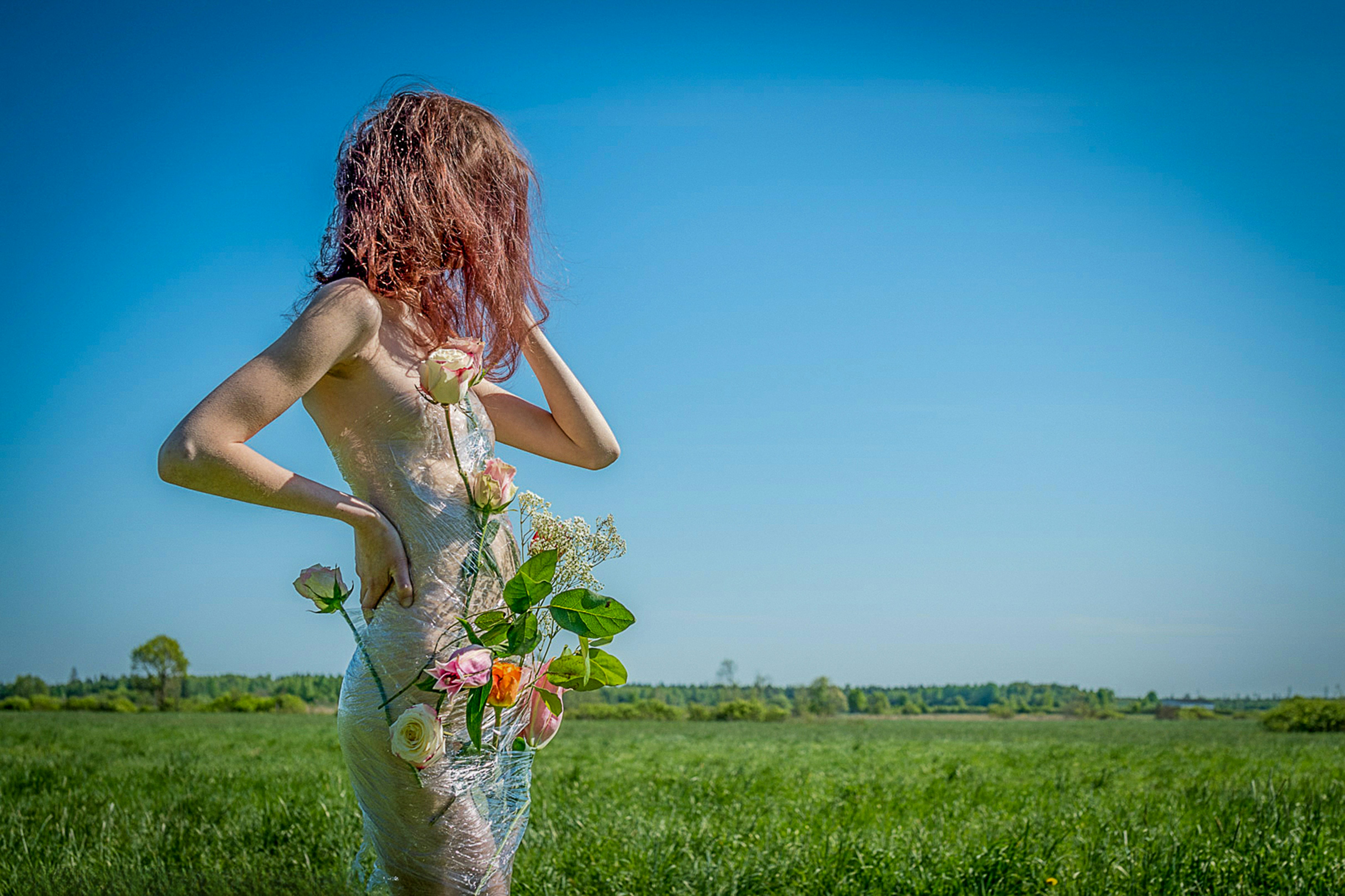 A creative conceptual fine art portrait of an anonymous young woman wrapped in clear plastic stretch film with fresh roses embedded inside, posing in a lush green meadow under a clear blue sky. 📸 Photo by me IG: @margoevardson