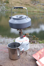 Camping kettle and stove with mug by a lake.