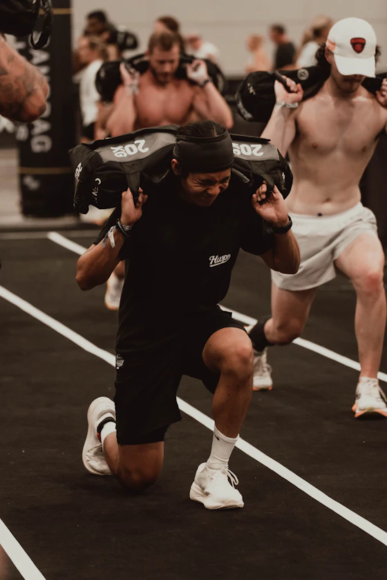 Atleta de camiseta do Hyrox executa afundos com sandbag de 20 kg nos ombros em pista de arena de competição, com outros atletas ao fundo