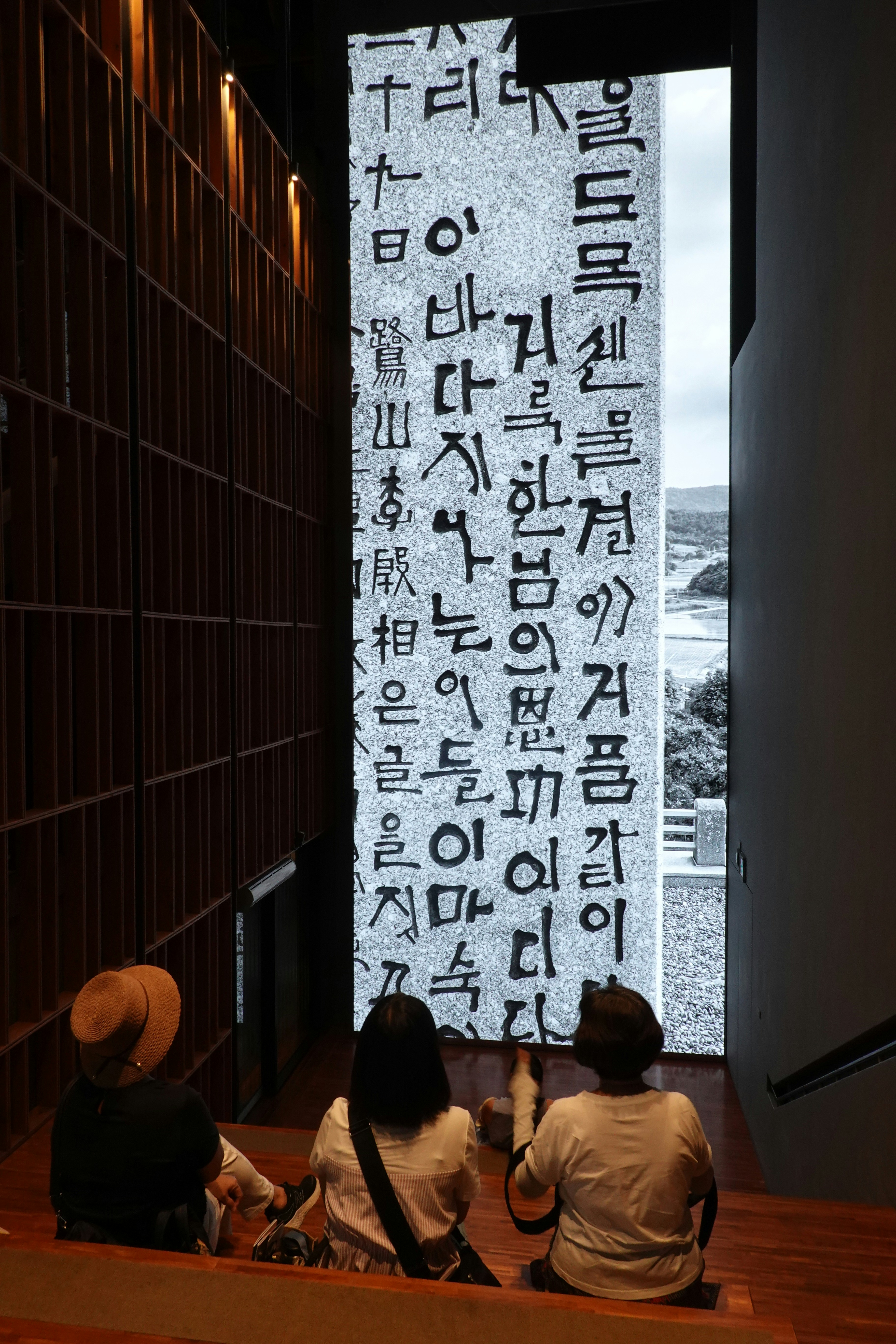 Three people admire a large screen displaying korean text.