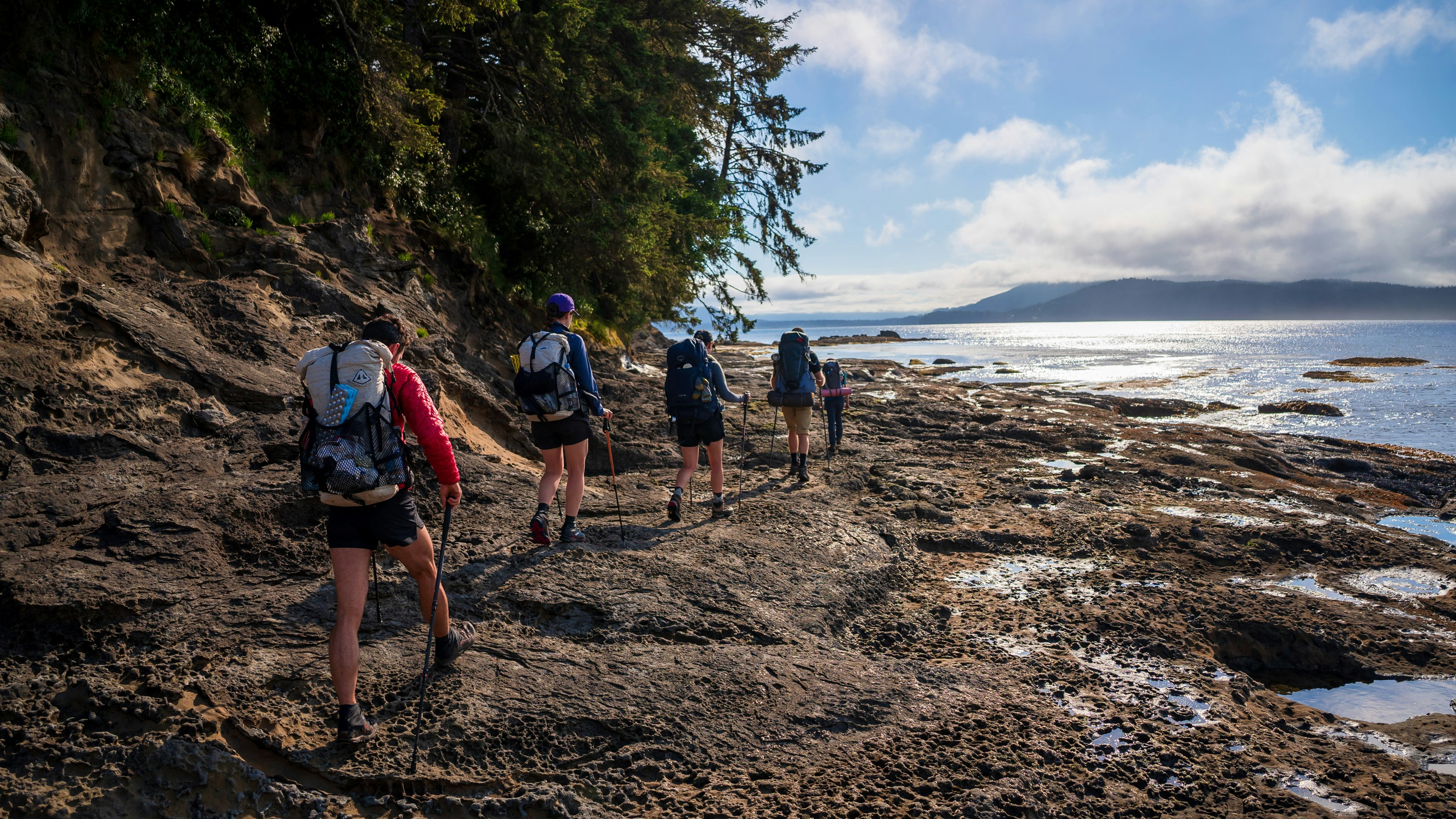 Four hikers walk along a rocky coastline beside the ocean.