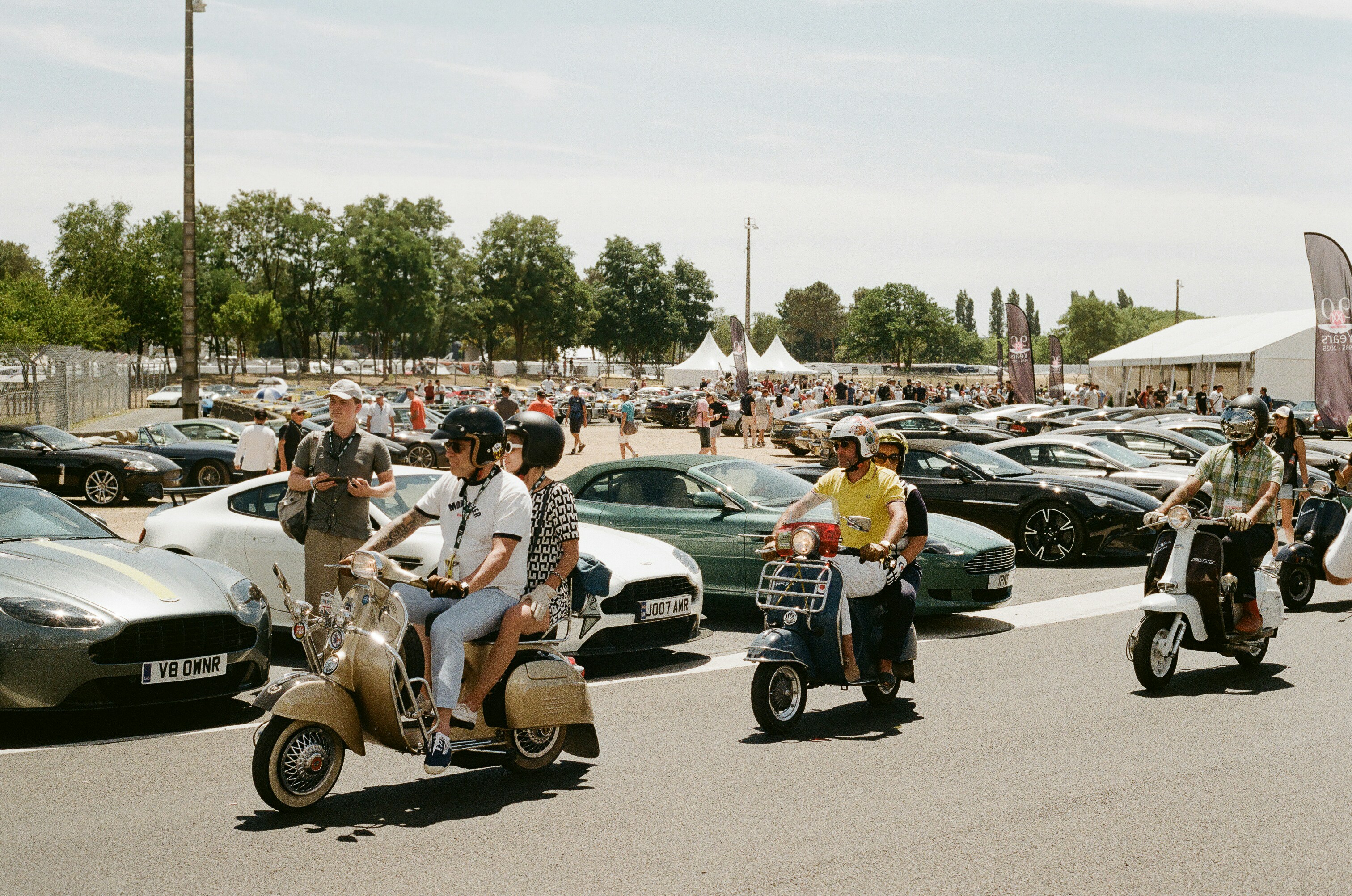 Riders on scooters passing parked luxury cars outdoors.