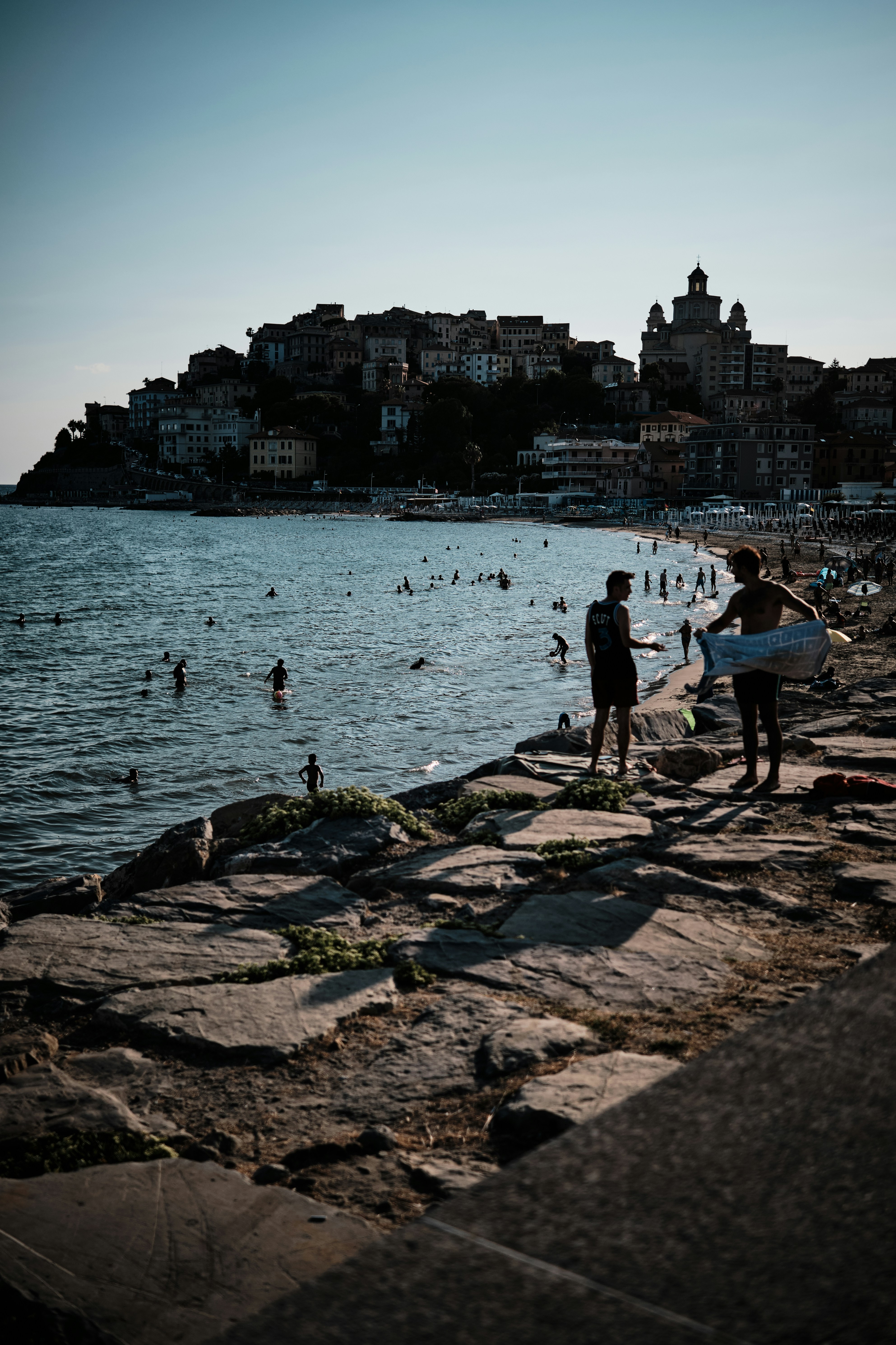Des surfeurs en silhouette sur des rochers surplombant une ville côtière animée.