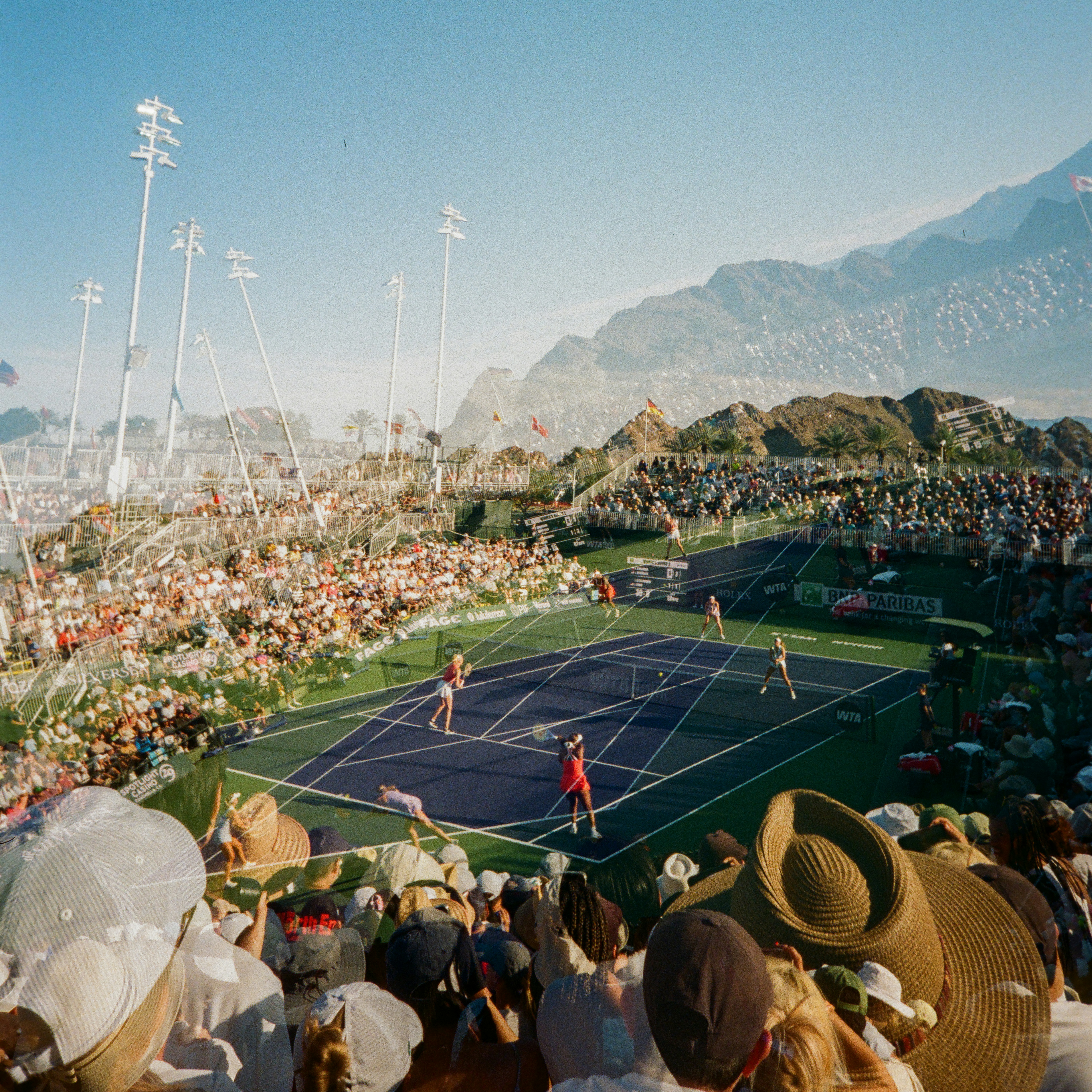 Pertandingan tenis luar ruangan dengan pemain, kerumunan, dan gunung di latar belakang.
