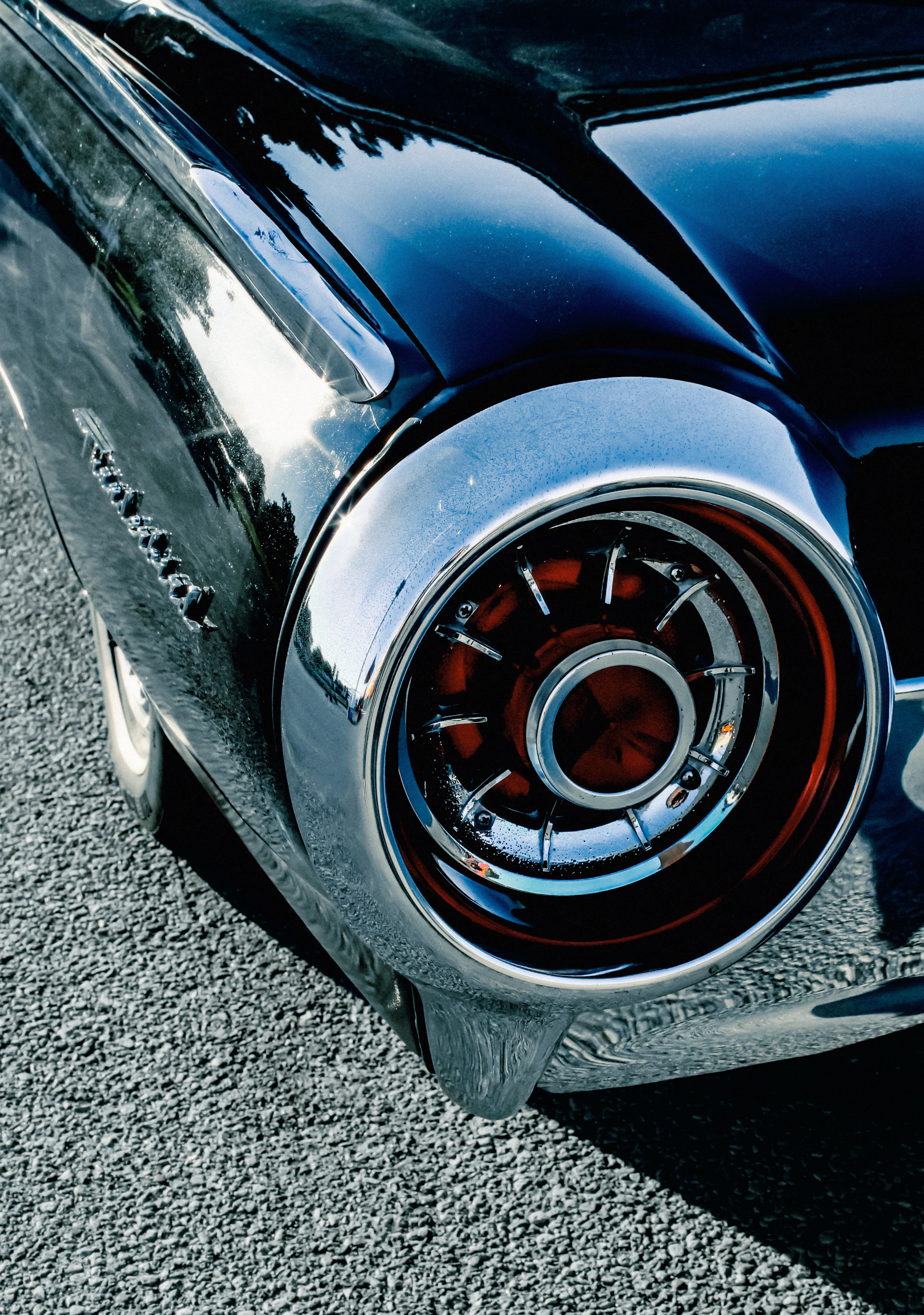Vintage car's gleaming chrome taillight and dark fender on asphalt.