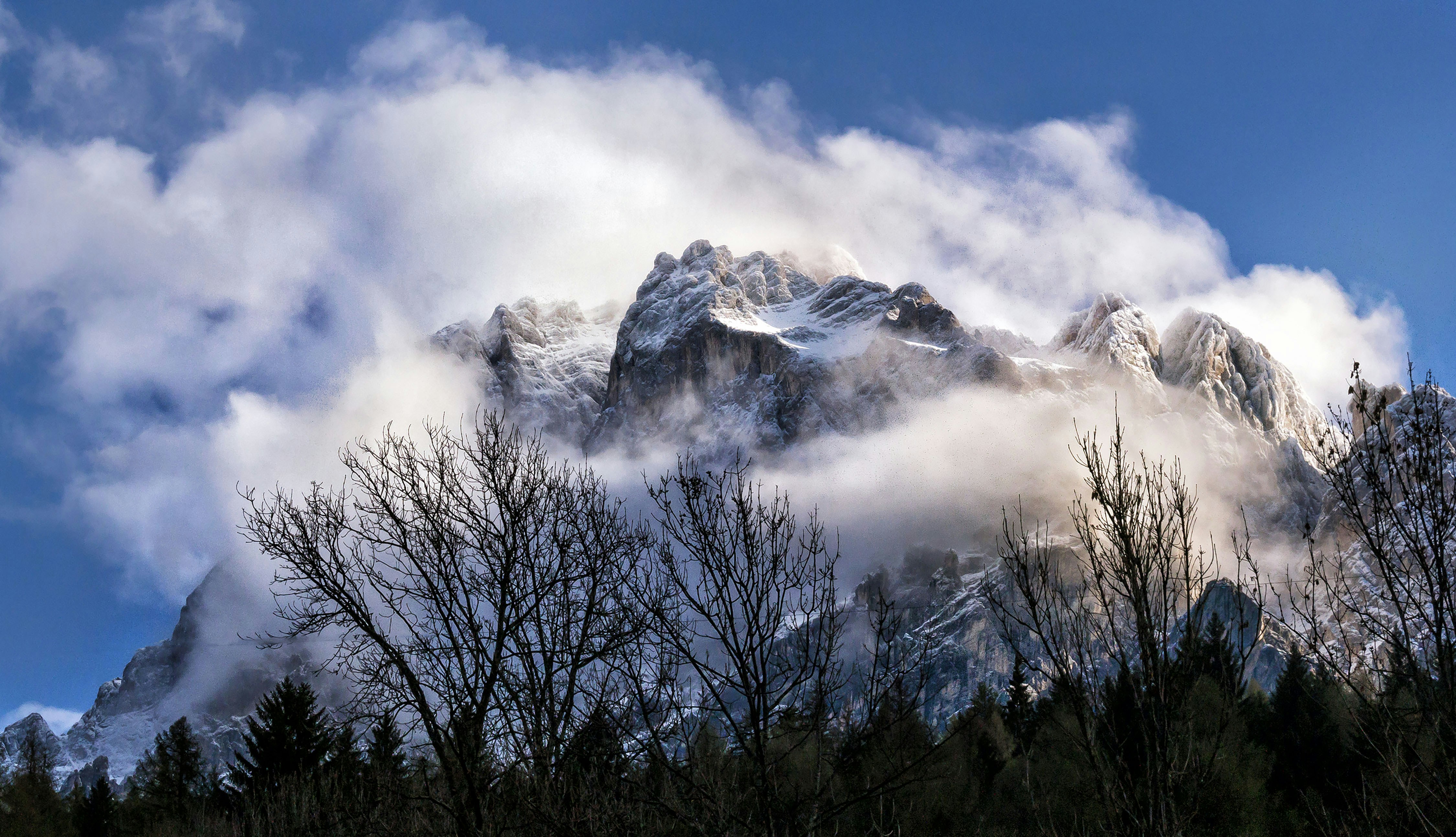 Montanhas cobertas de neve envoltas em nuvens, com uma floresta abaixo.
