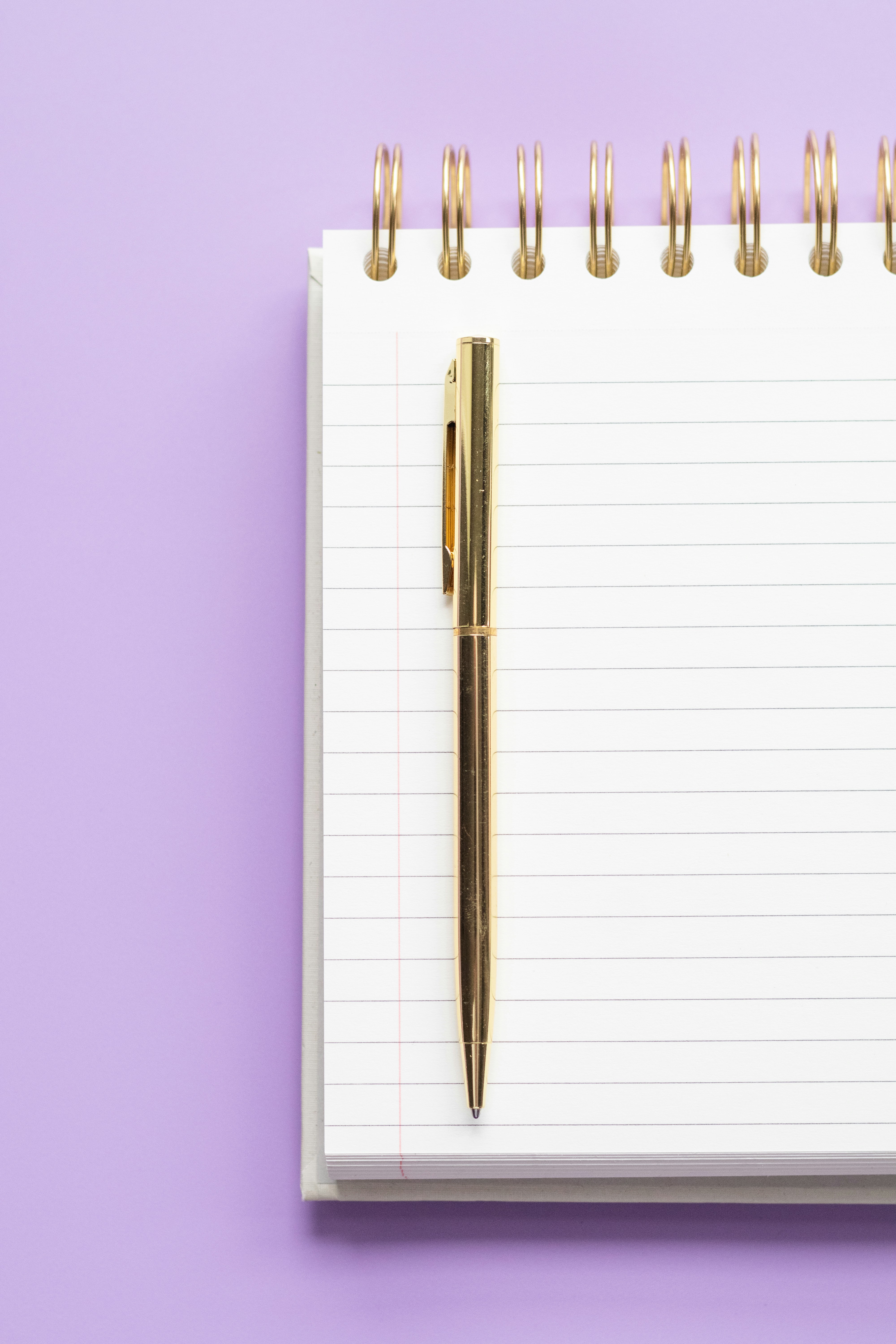 White spiral notebook and gold pen on purple background.