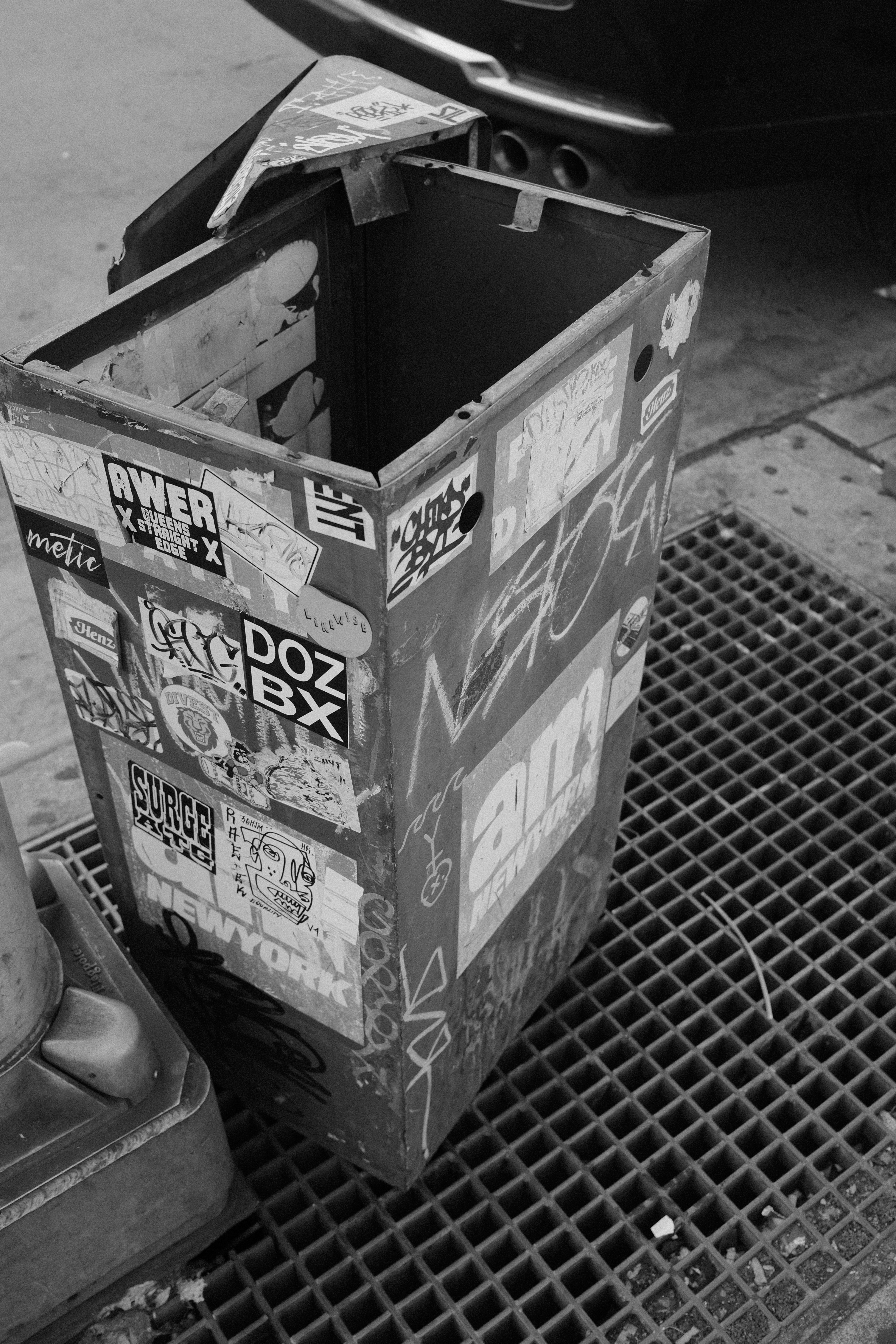 A sticker-covered trash can sits next to a metal grate.