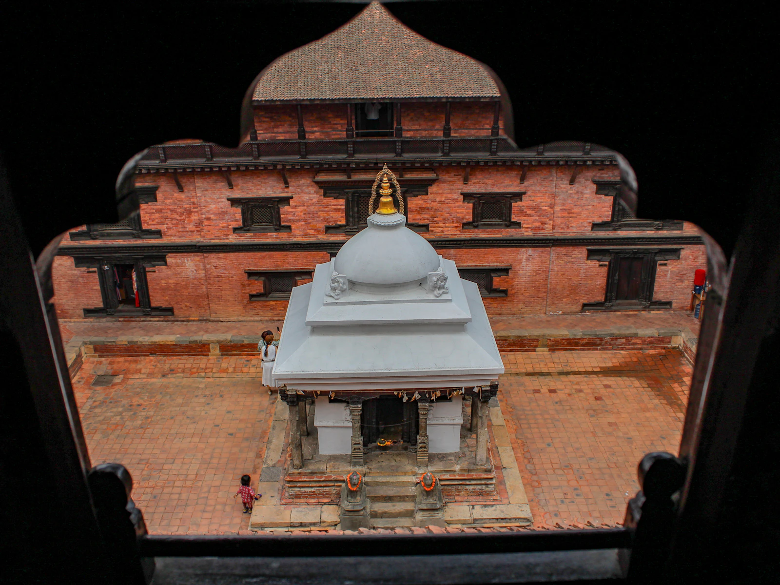 A Patan Durbar Square courtyard of red brick and carved windows, seen from above through a dark wooden window frame