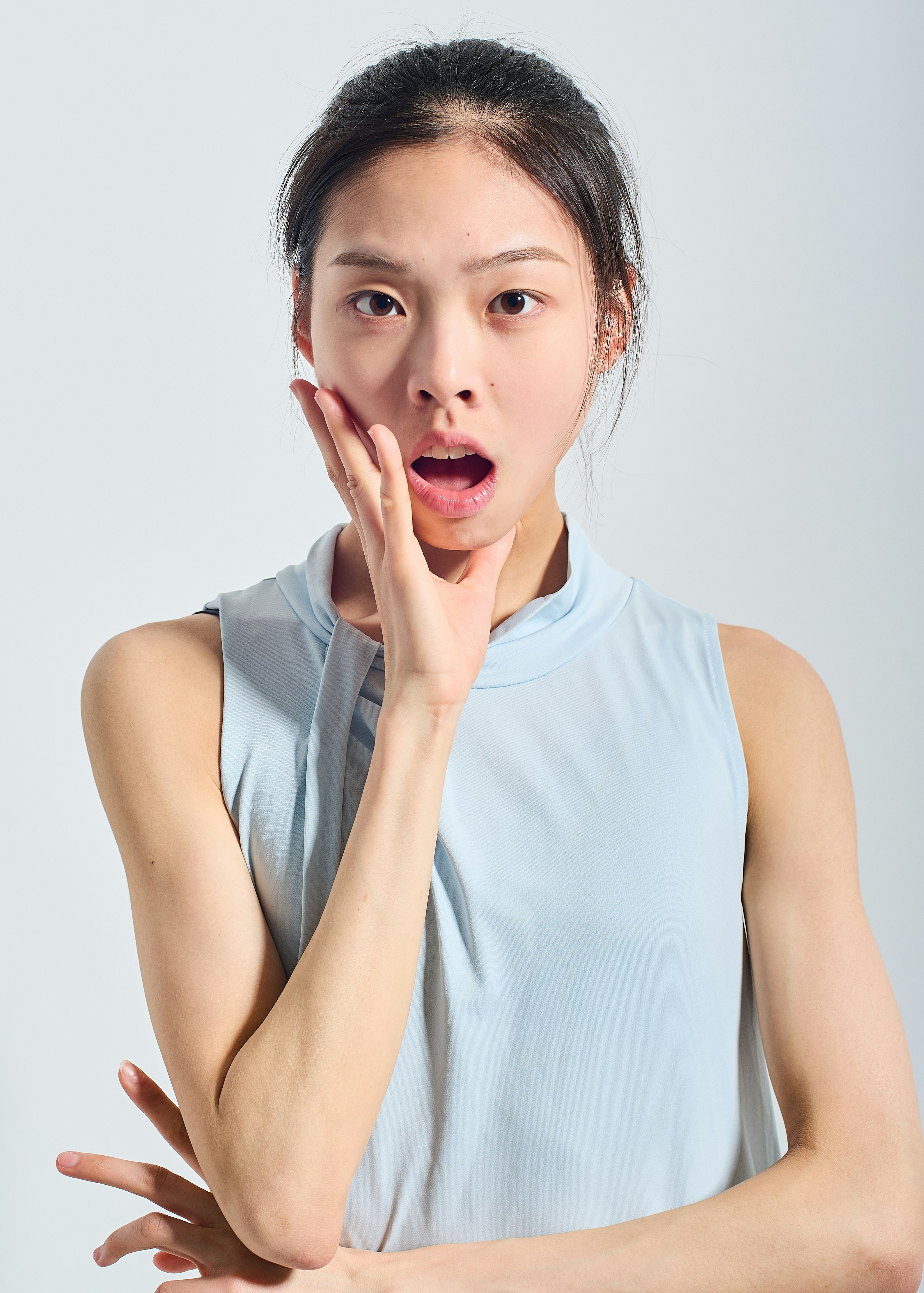 Close-up portrait of a surprised woman with hand near her mouth against a pale background. A logo-free studio shot focused on facial expression and upper torso.
